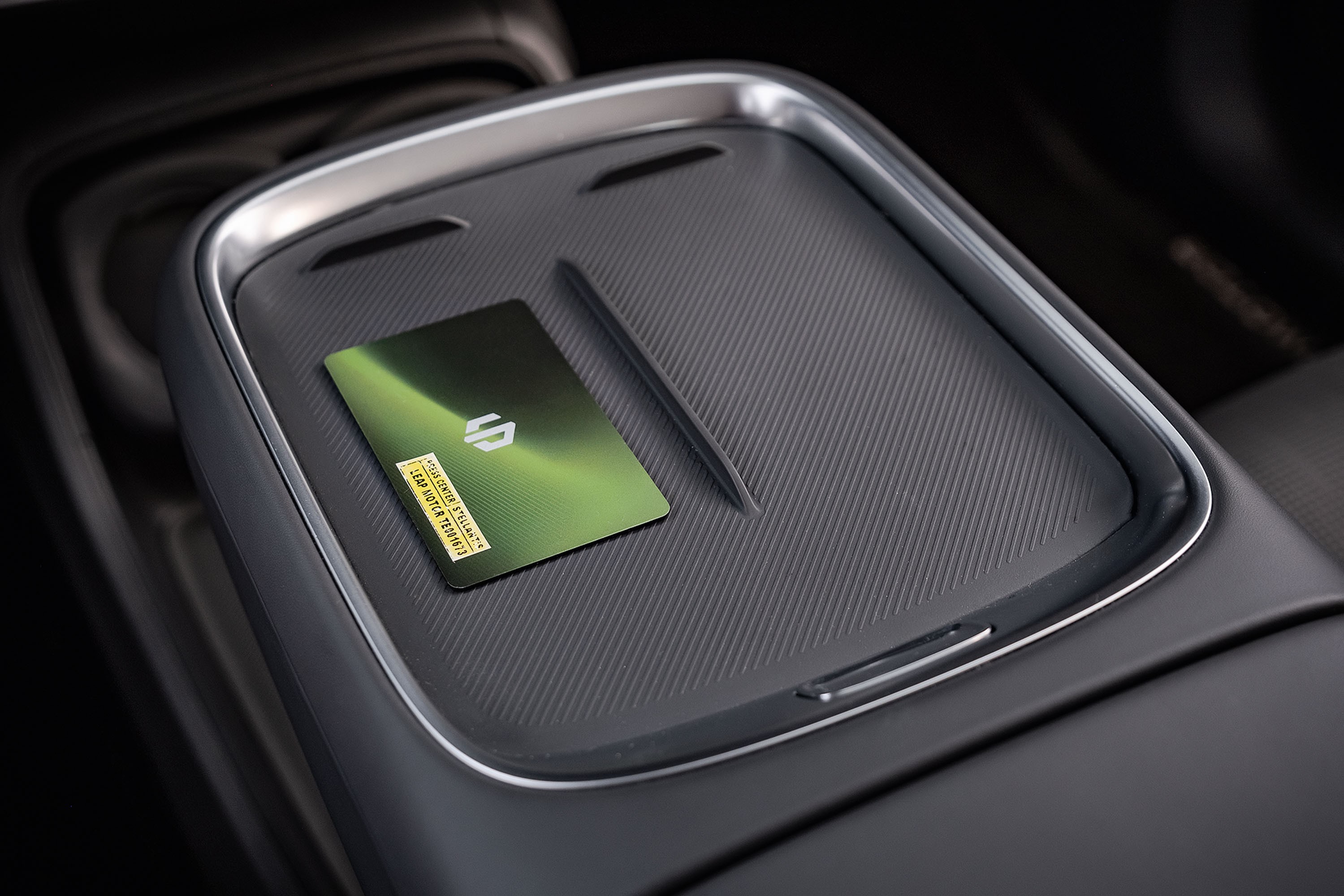 Cartão verde metálico com logo branco e adesivo amarelo sobre console central cinza escuro texturizado de carro, com detalhes prateados e uma caneta preta fina ao lado
