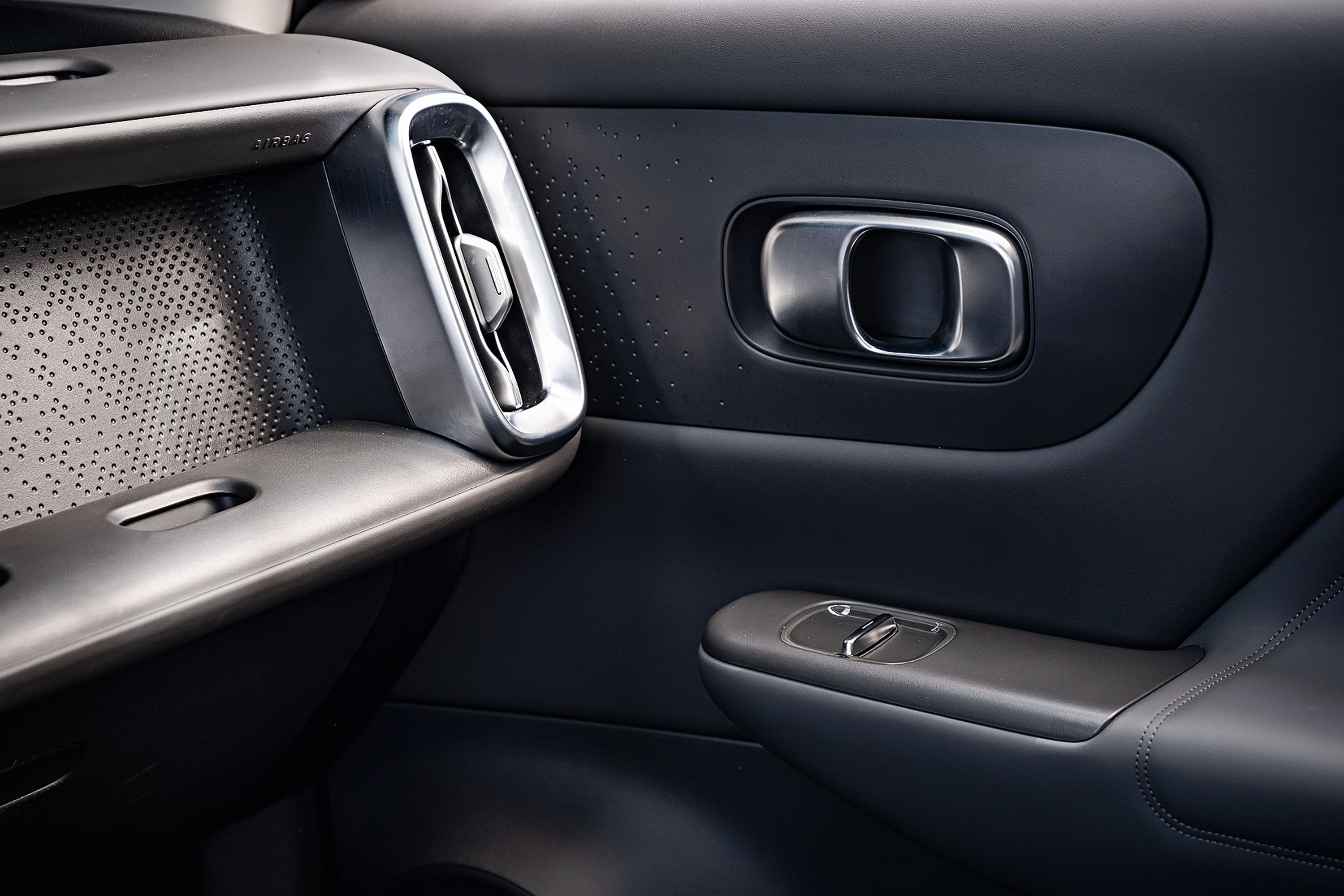 Interior de carro moderno com detalhes em cinza escuro e prata. À esquerda, painel texturizado com pequenos furos e saída de ar vertical cromada. À direita, maçaneta da porta em metal escovado e, abaixo, controles dos vidros elétricos com acabamento em cinza e prata