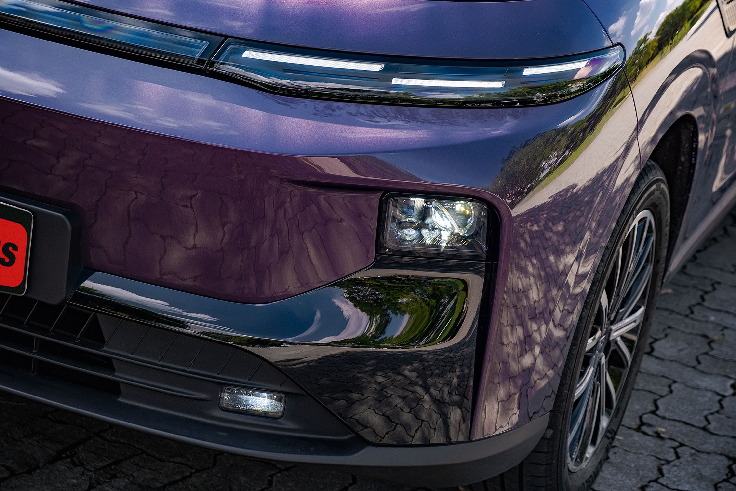 Parte frontal de um carro roxo metálico, com faróis de LED horizontais e um farol principal quadrado aceso. A superfície reflete o céu e árvores, e a roda com aros prateados é visível à direita. O veículo está sobre um piso de paralelepípedos