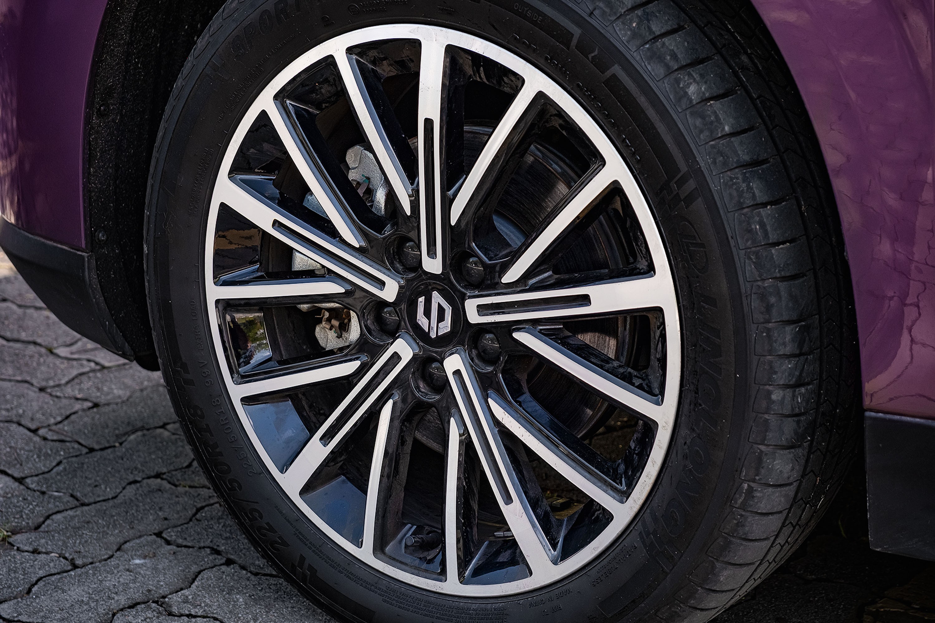Roda de carro com aro preto e detalhes prateados, pneu com sulcos visíveis, parte da lataria roxa e chão de paralelepípedos