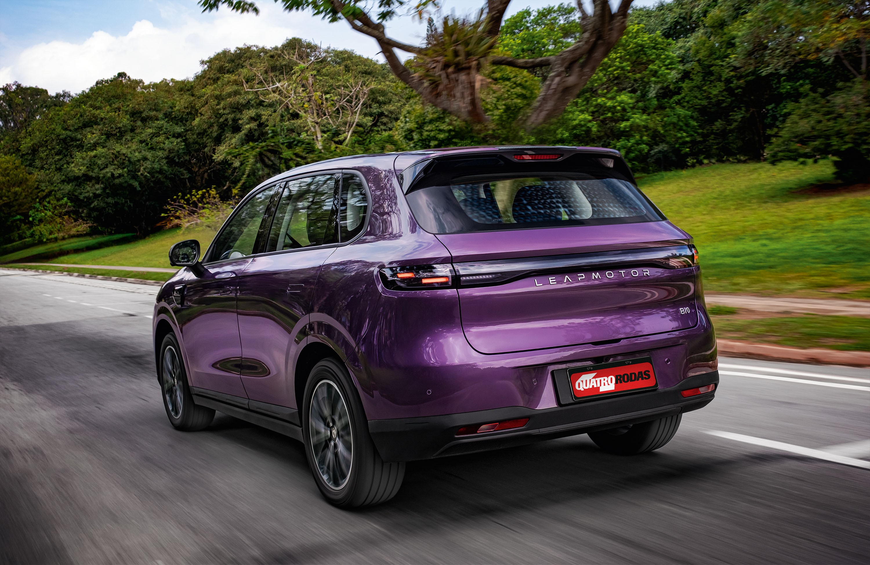 SUV Leapmotor C10 roxo metálico em movimento, visto por trás, em uma estrada asfaltada com árvores verdes ao fundo e céu azul com nuvens