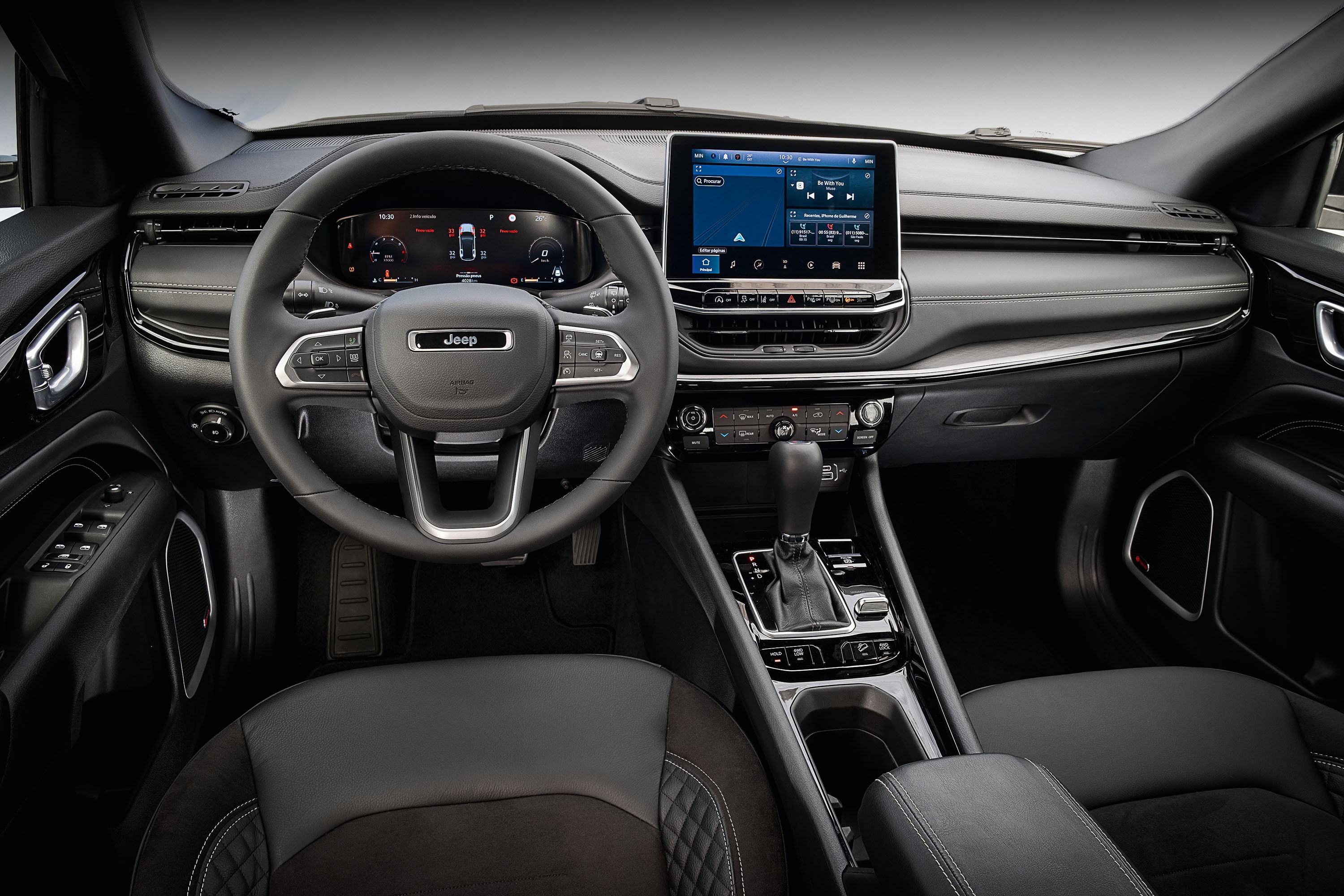 Interior de um carro moderno em tons escuros, com volante multifuncional da marca Jeep, painel digital e tela multimídia central exibindo um reprodutor de música. O console central possui câmbio automático e botões de controle, e o banco do motorista é de couro preto com costura em losango