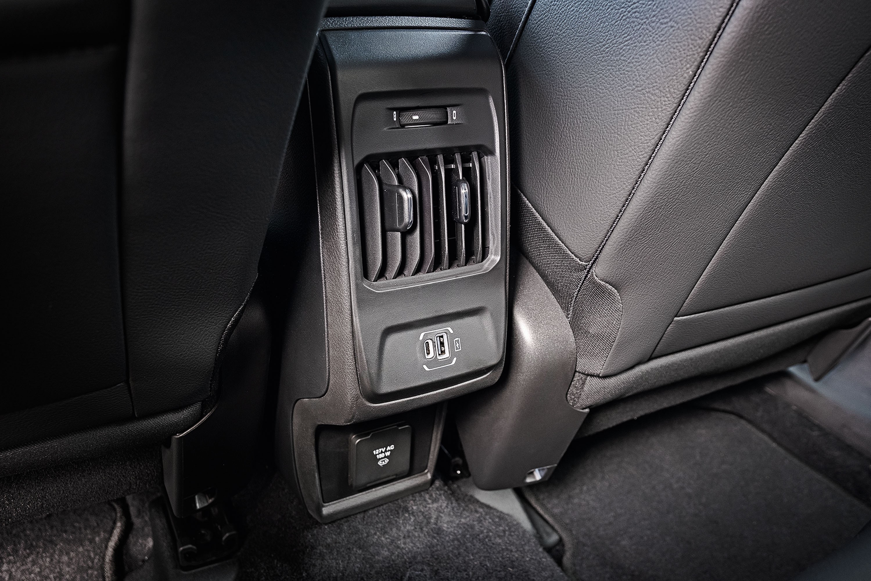 Painel traseiro de um carro com saídas de ar, duas portas USB e uma tomada de 12V, entre dois bancos de couro preto