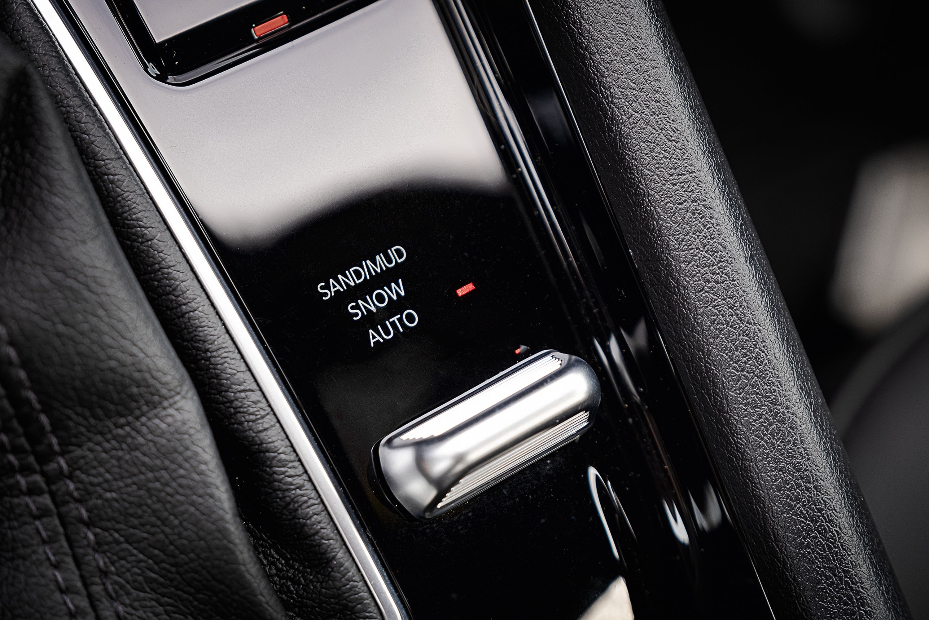 Painel de controle de carro com seletor de modo de condução. As opções visíveis são SAND/MUD, SNOW e AUTO, com um indicador vermelho ao lado de SNOW. Um botão cromado alongado está abaixo das opções, e o painel é preto brilhante, ladeado por estofamento de couro preto texturizado