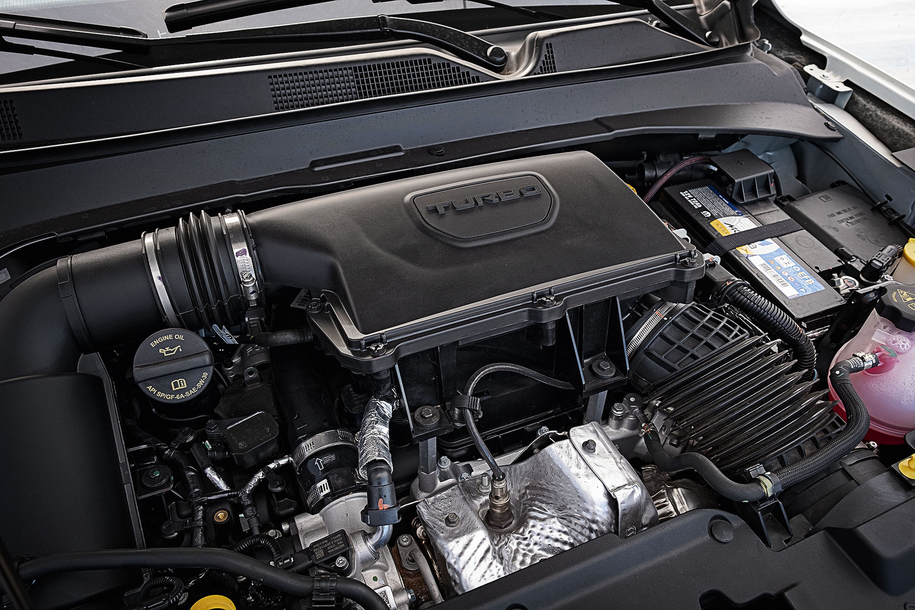 Vista superior do motor de um carro, com a tampa preta do motor exibindo a palavra TURBO em relevo. À esquerda, o bocal de óleo com o símbolo de uma almotolia. À direita, a bateria do veículo e o reservatório de fluido rosa. Diversos tubos, fios e componentes metálicos prateados estão visíveis, com o para-brisa e a grade de ventilação do carro ao fundo