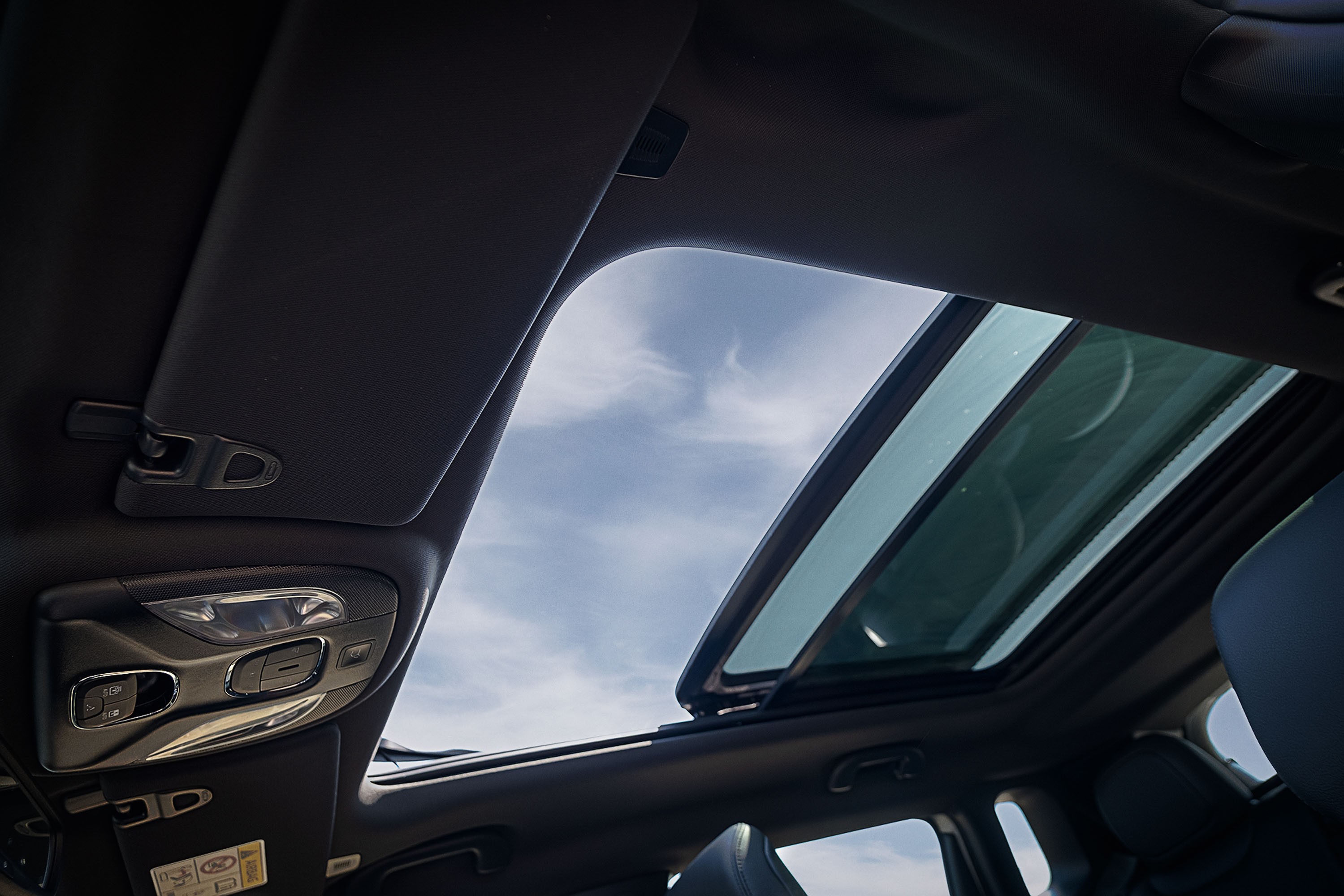 Vista interna do teto de um carro com teto solar panorâmico aberto, revelando um céu azul com nuvens esparsas. O interior é escuro, com detalhes do console de luzes e para-sol visíveis