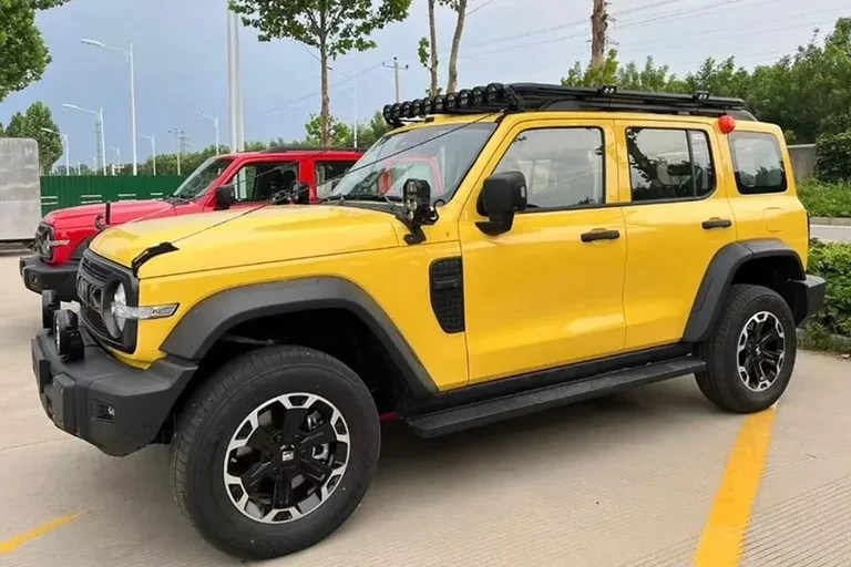 Jipe amarelo com teto solar, bagageiro e faróis auxiliares no teto, estacionado em uma rua com outro jipe vermelho ao fundo