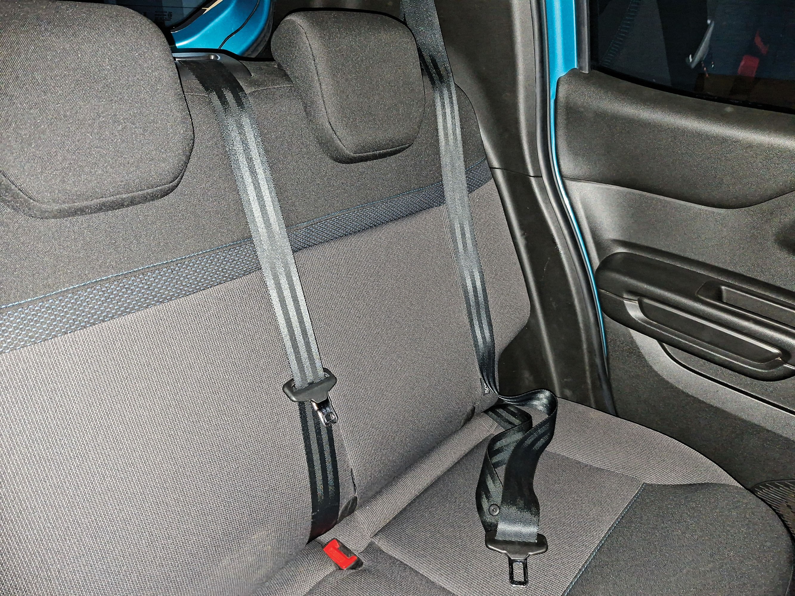Bancos traseiros de carro com estofamento cinza e detalhes em preto, com dois cintos de segurança pretos visíveis, um afivelado e outro solto, e parte da porta azul do veículo à direita