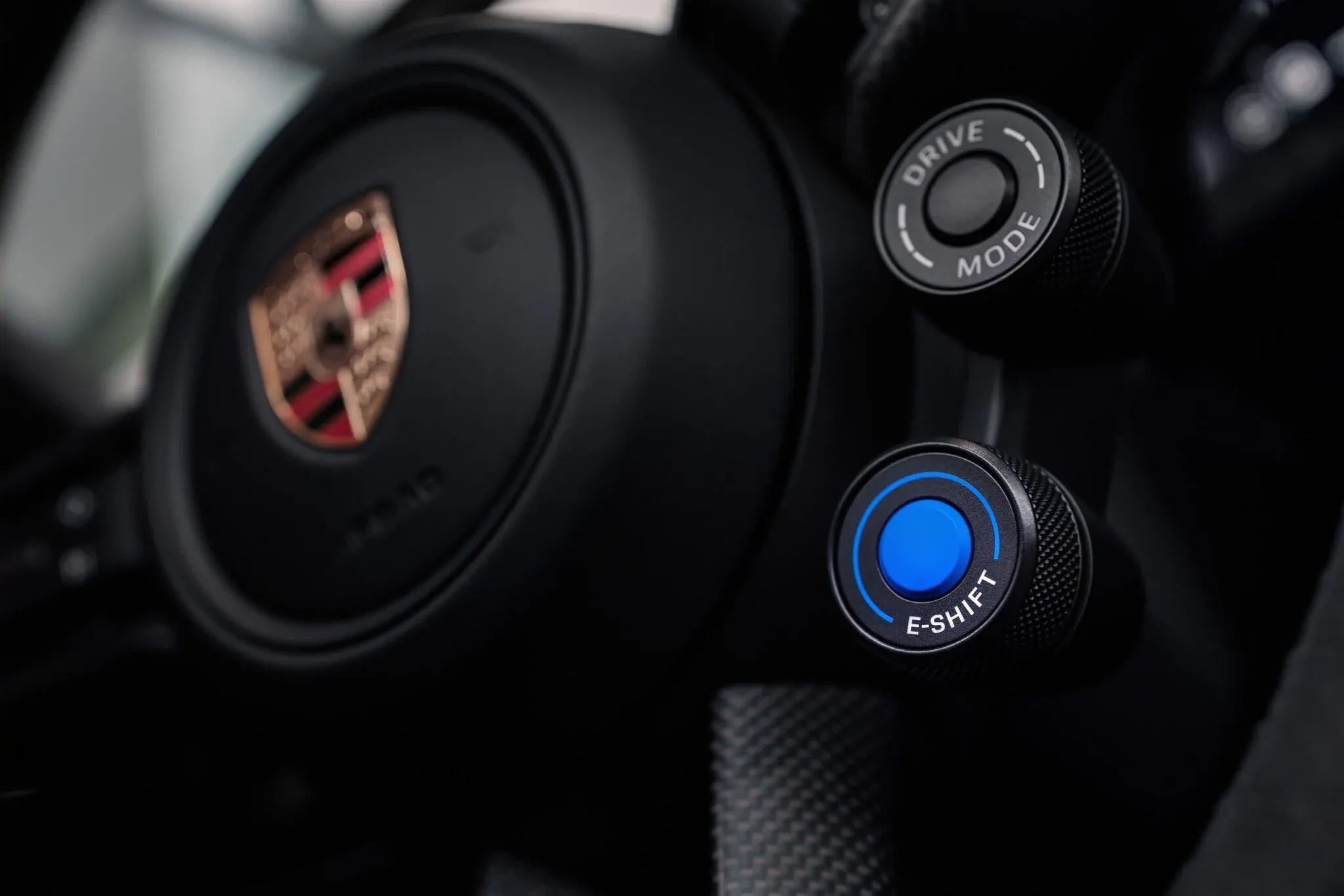 Detalhe do volante de um carro Porsche, com o emblema da marca no centro e dois botões giratórios pretos à direita, um para DRIVE MODE e outro para E-SHIFT com um círculo azul brilhante
