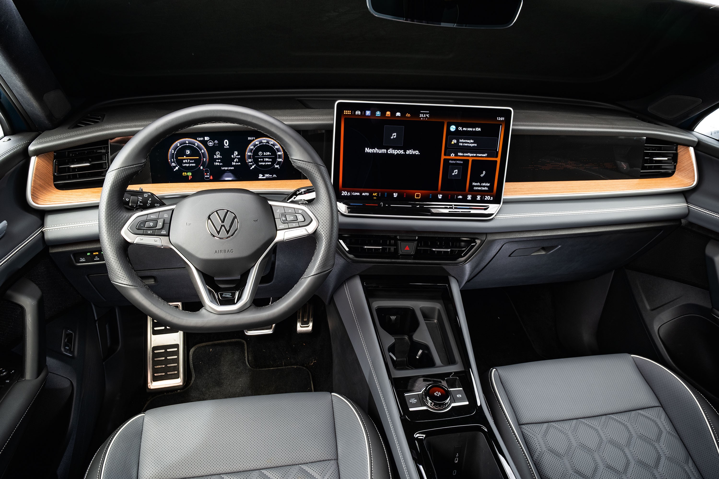 Interior de um carro moderno, com volante multifuncional da Volkswagen e painel digital. À direita, uma tela multimídia exibe Nenhum dispositivo ativo. Detalhes em madeira clara no painel e estofamento cinza com costuras brancas nos bancos. Pedais metálicos e console central com porta-copos e botões de controle.