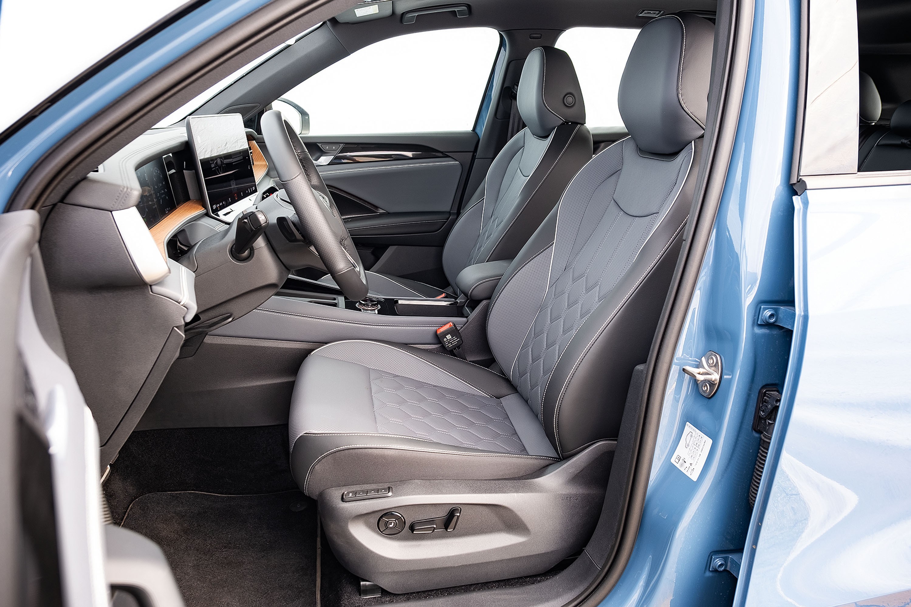 Interior de um carro azul, mostrando o banco do motorista e do passageiro da frente. Os bancos são cinza e preto com costura branca e detalhes em losango. O volante e o painel digital são visíveis, com um acabamento em madeira clara no painel. O chão é forrado com carpete escuro. A porta do motorista está aberta, revelando a parte interna da lataria azul