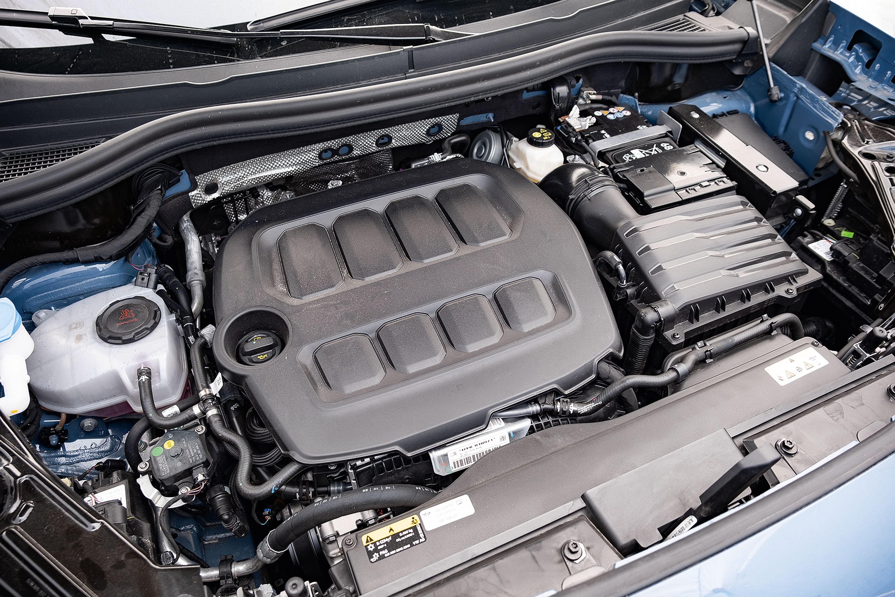 Vista superior do motor de um carro, com a tampa do motor preta em destaque no centro, cercada por mangueiras, reservatórios e componentes metálicos e plásticos, em um compartimento azul