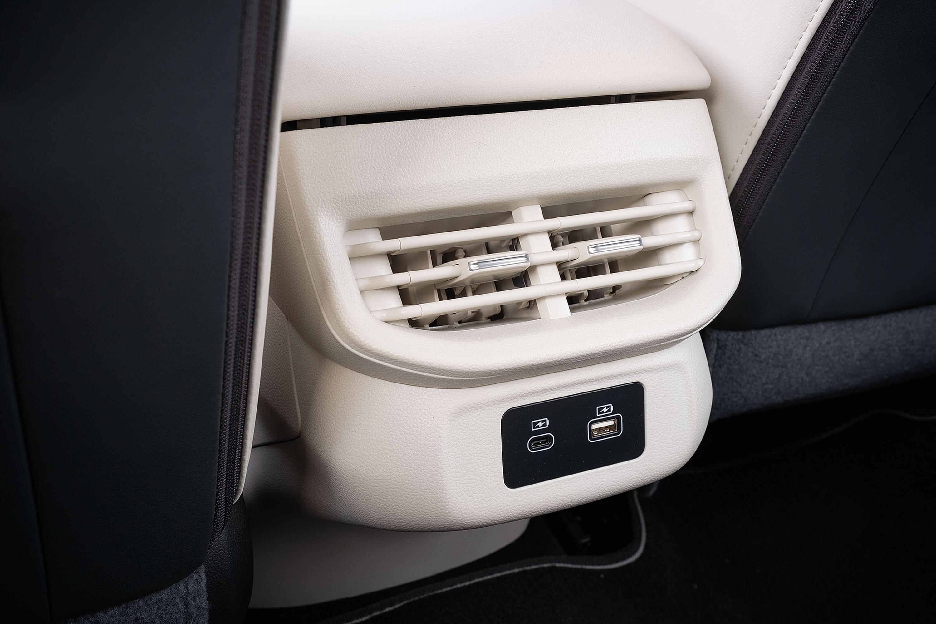 Saída de ar-condicionado e duas portas USB, uma tipo C e uma tipo A, na parte traseira do console central de um carro, com estofamento claro e carpete escuro