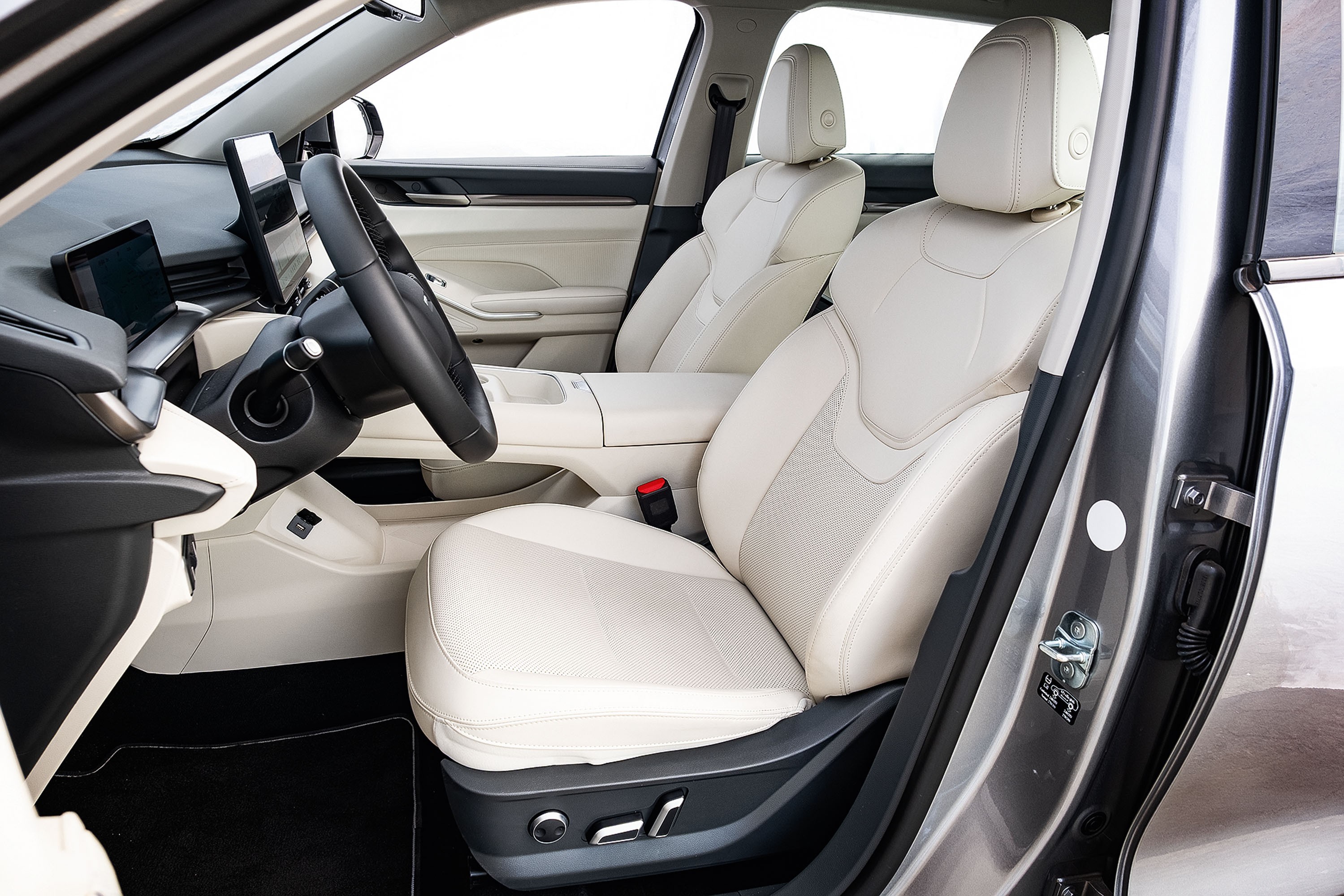 Interior de carro moderno com bancos de couro branco, volante escuro e painel com telas digitais. O banco do motorista possui ajustes elétricos visíveis na lateral. O ambiente é claro, com janelas abertas revelando um fundo branco