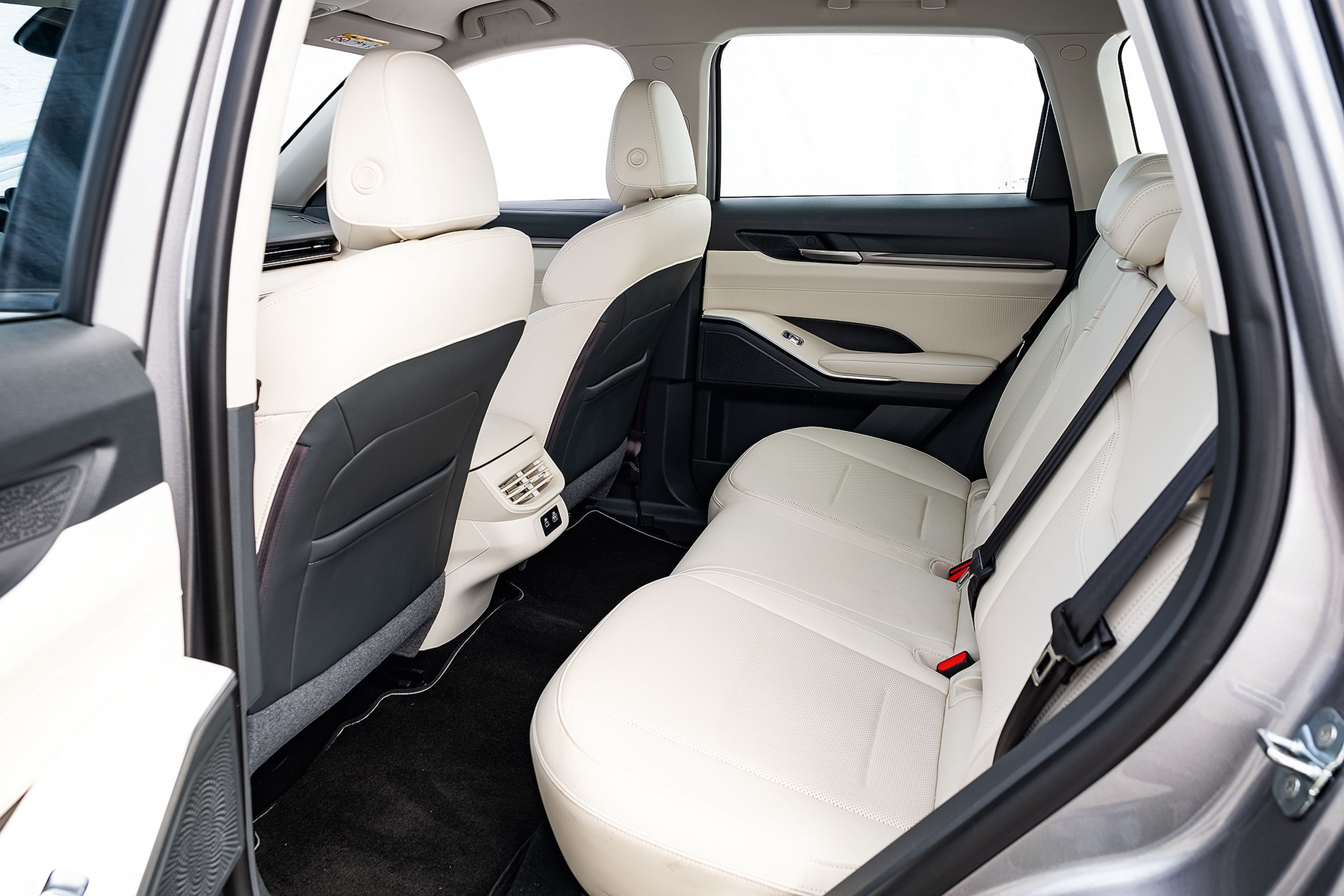 Interior de carro com bancos de couro creme e detalhes em preto, incluindo encostos e laterais das portas. O banco traseiro tem cintos de segurança pretos e fivelas vermelhas, com saídas de ar no console central. O carpete é preto, e as janelas mostram um fundo branco