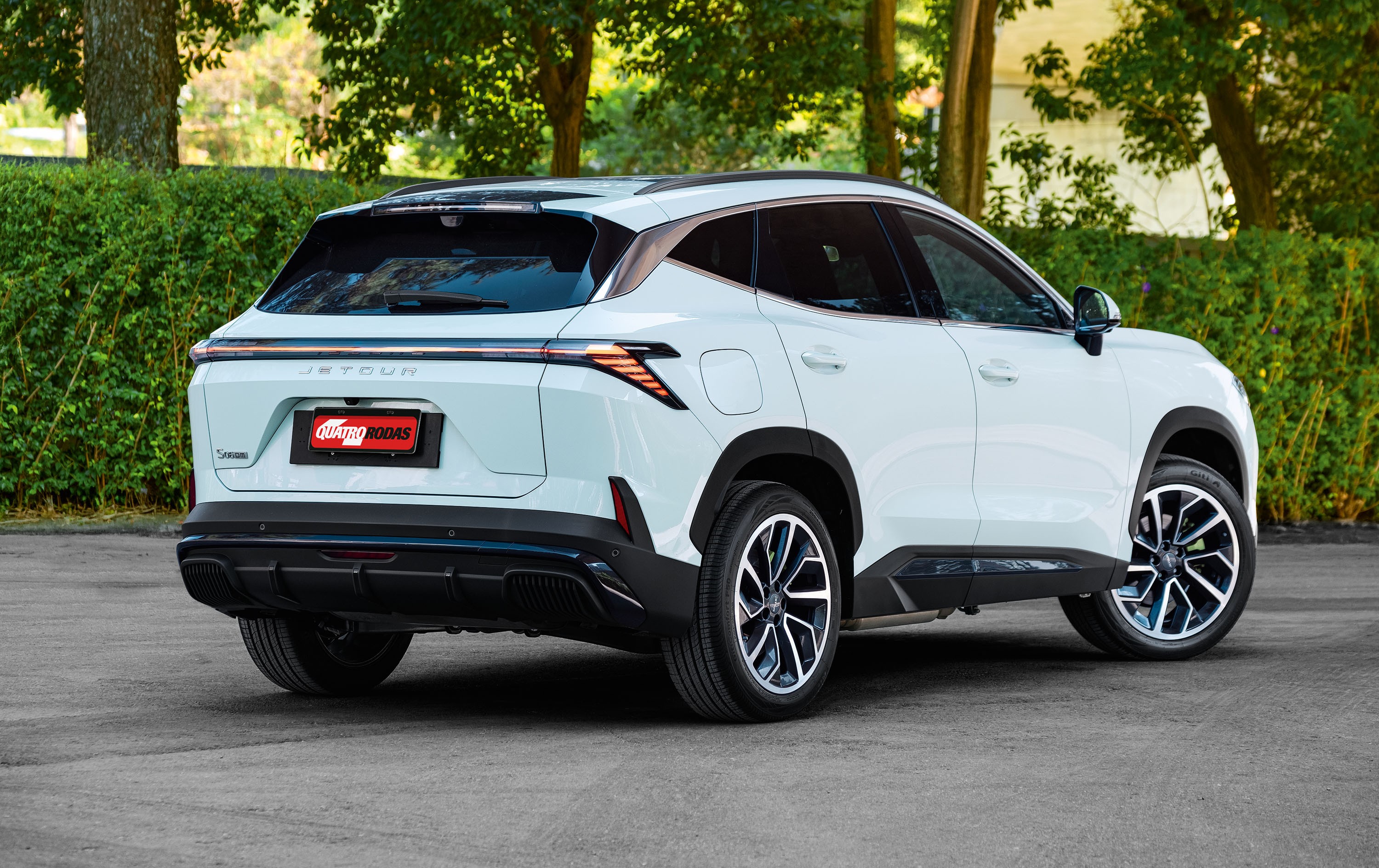 SUV Jetour branco, visto de traseira e lateral, estacionado em asfalto cinza com vegetação verde ao fundo. A placa traseira vermelha exibe QUATRO RODAS