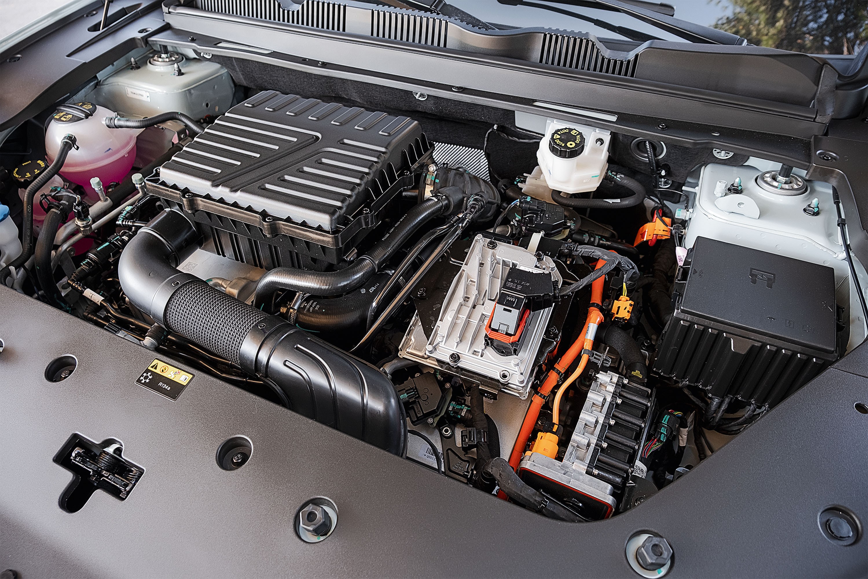 Motor de carro híbrido com componentes elétricos e mecânicos. Destacam-se cabos laranja, reservatórios de fluidos, caixa de fusíveis preta e um grande filtro de ar.