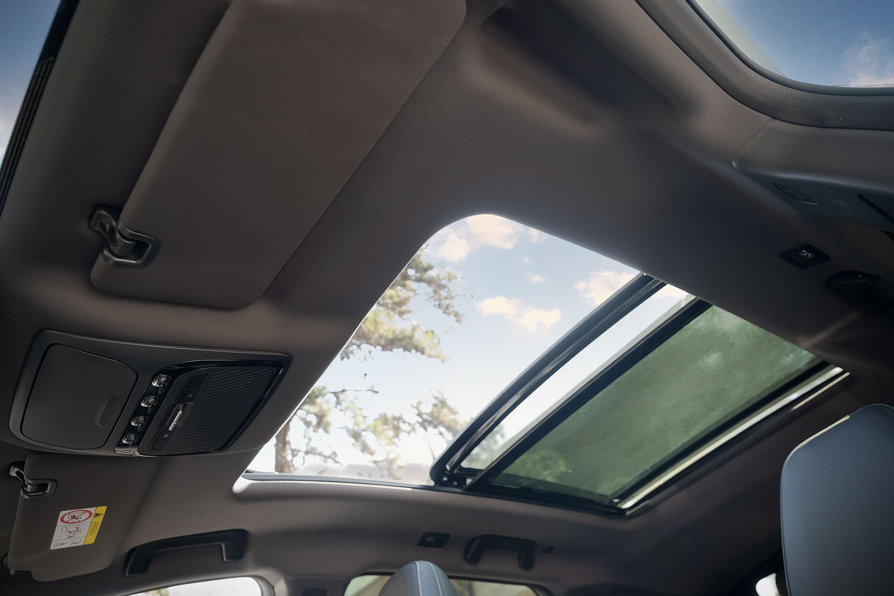 Interior de um carro com teto solar panorâmico aberto, mostrando o céu azul com nuvens e árvores ao fundo. O forro do teto é cinza, com para-sol e console de luzes e botões