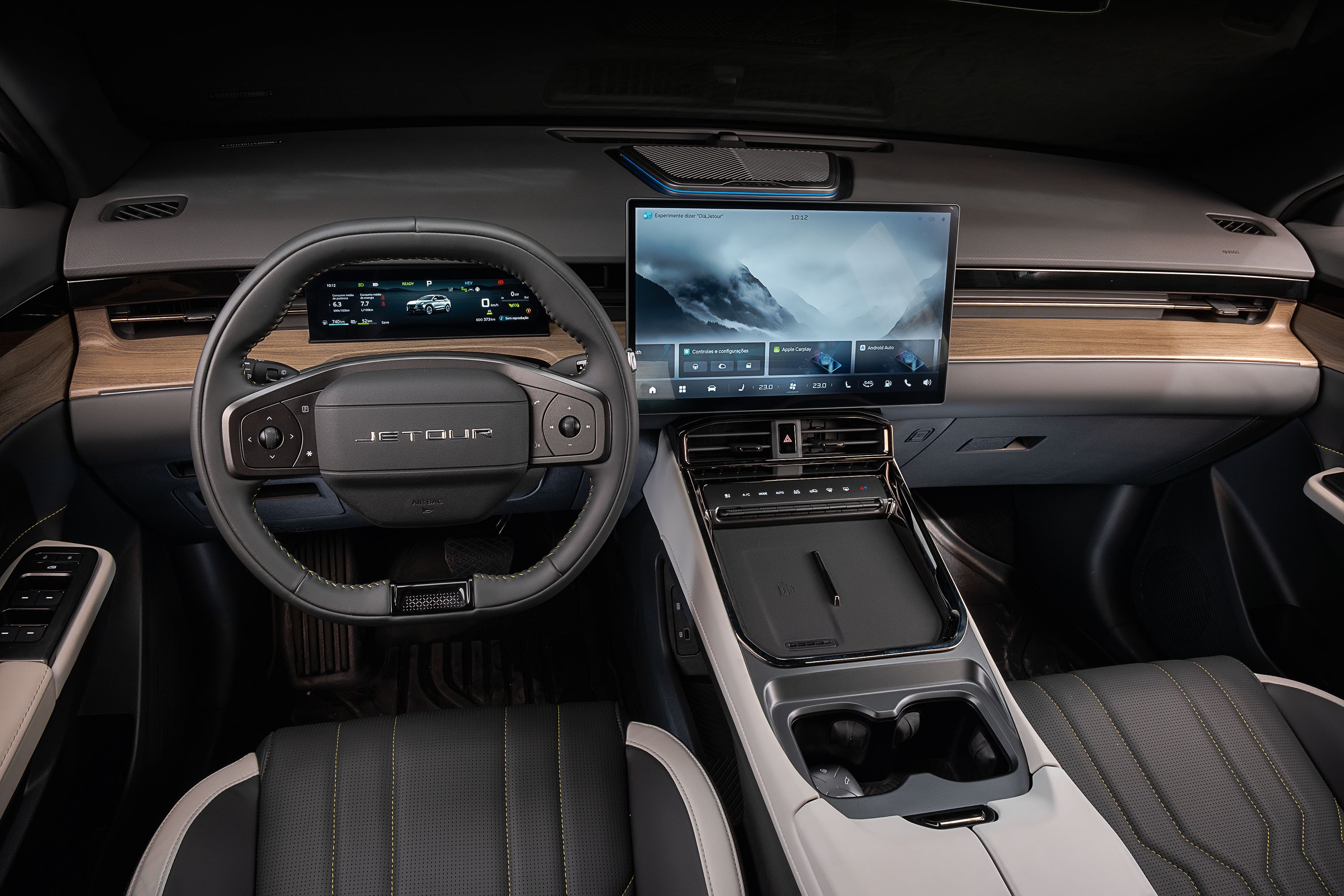 Interior moderno de um carro, com volante preto e o nome JETOUR no centro. O painel possui uma tela digital exibindo um carro e outra tela maior no centro com uma paisagem montanhosa e opções de menu. Detalhes em madeira clara e acabamentos cinza escuro e prata complementam o design, com bancos de couro preto e costura amarela visíveis no canto inferior direito