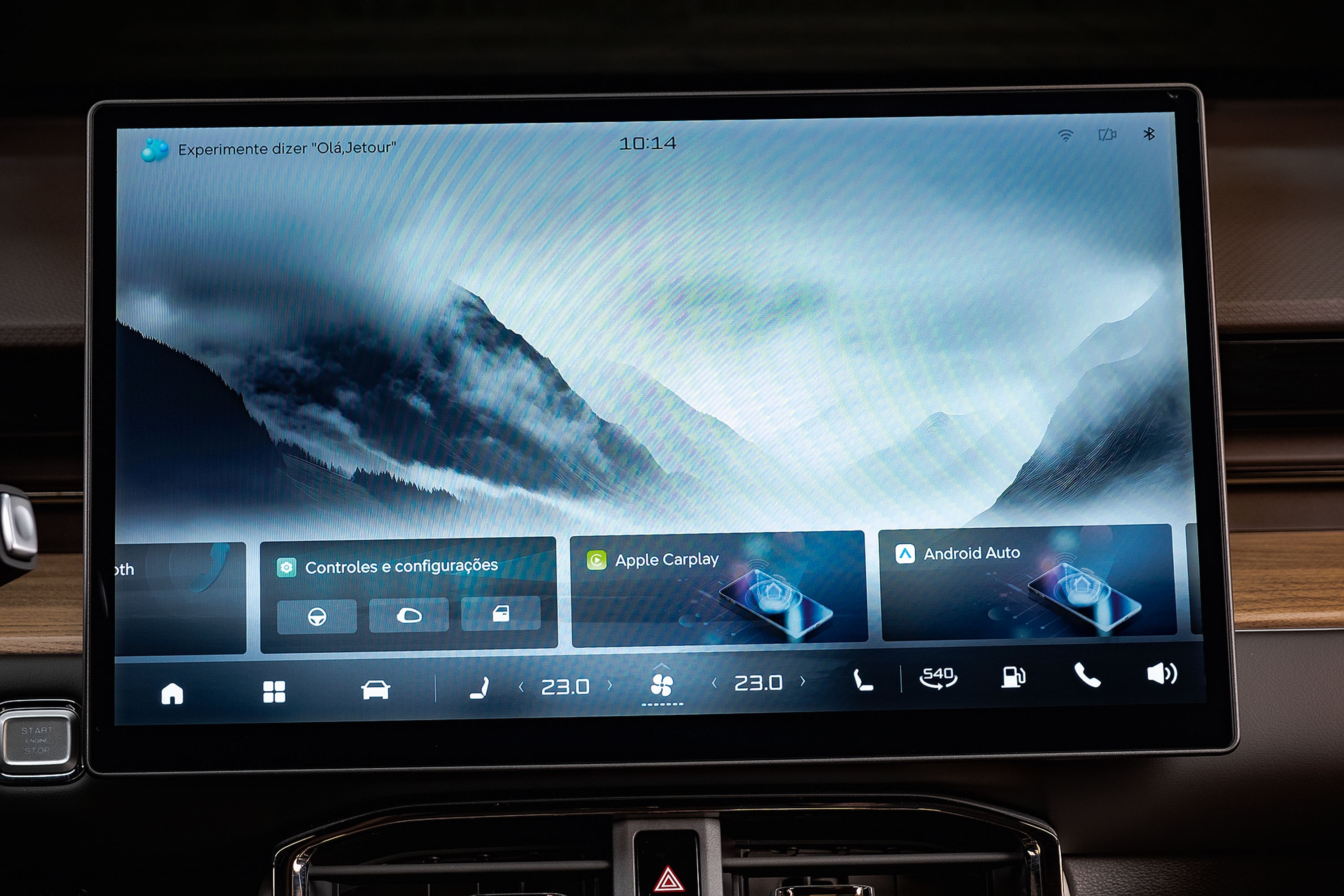 Tela multimídia de carro exibindo um papel de parede de montanhas nebulosas. Na parte inferior, ícones de navegação, controles do veículo, Apple CarPlay e Android Auto. No topo, a hora 10:14 e a sugestão Experimente dizer Olá Jetour.