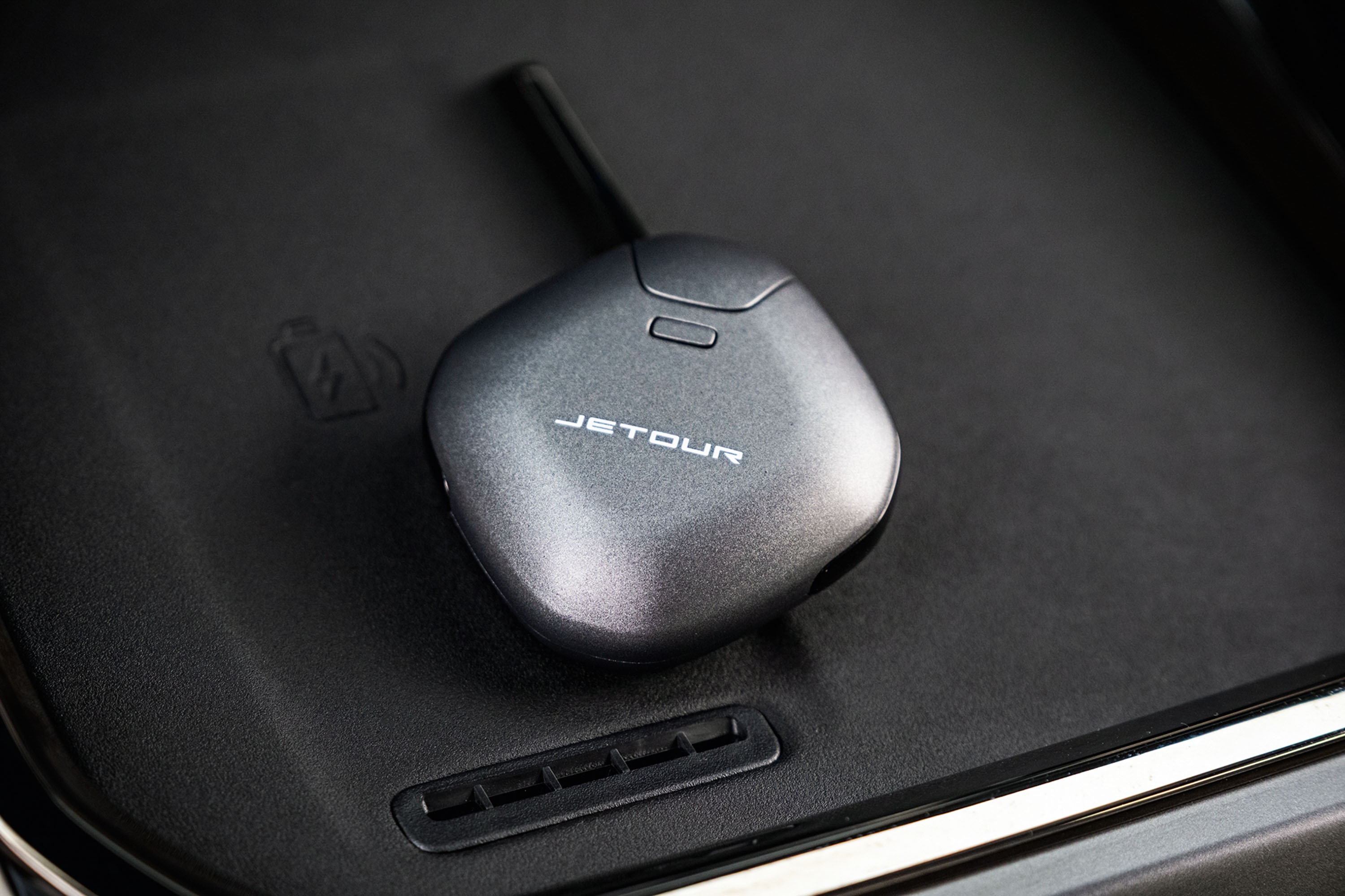Chave de carro Jetour cinza-chumbo com antena preta, repousando sobre um console preto texturizado de veículo, com um ícone de carregamento sem fio em relevo
