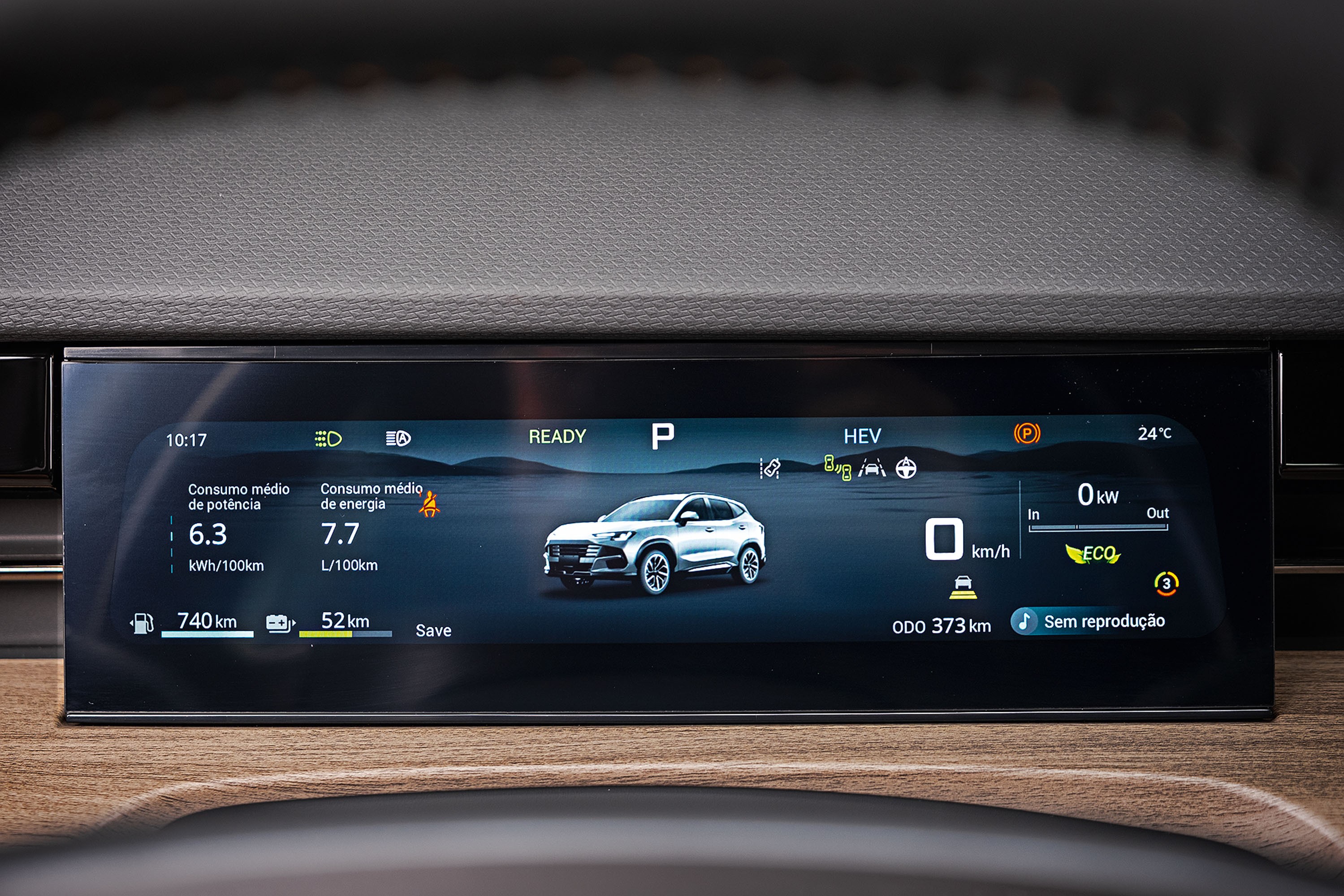 Painel digital de carro exibindo informações de consumo, autonomia e um SUV branco ao centro. O consumo de potência é 6.3 kWh/100km e o de energia 7.7 L/100km. A autonomia é de 740 km de combustível e 52 km de bateria. O carro está em modo P (estacionado) e HEV (híbrido), com temperatura de 24°C. O odômetro marca 000 373 km e não há reprodução de mídia. O fundo é uma paisagem montanhosa azul escura