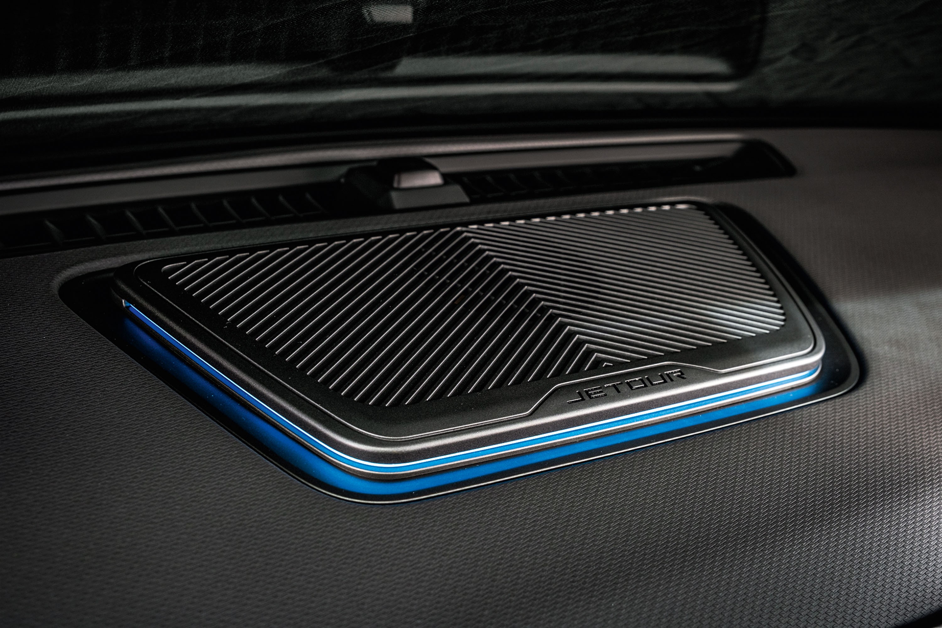 Detalhe do interior de um carro, mostrando um alto-falante retangular com grade metálica e iluminação ambiente azul ao redor, sobre um painel escuro texturizado. A marca JETOUR está gravada na borda do alto-falante