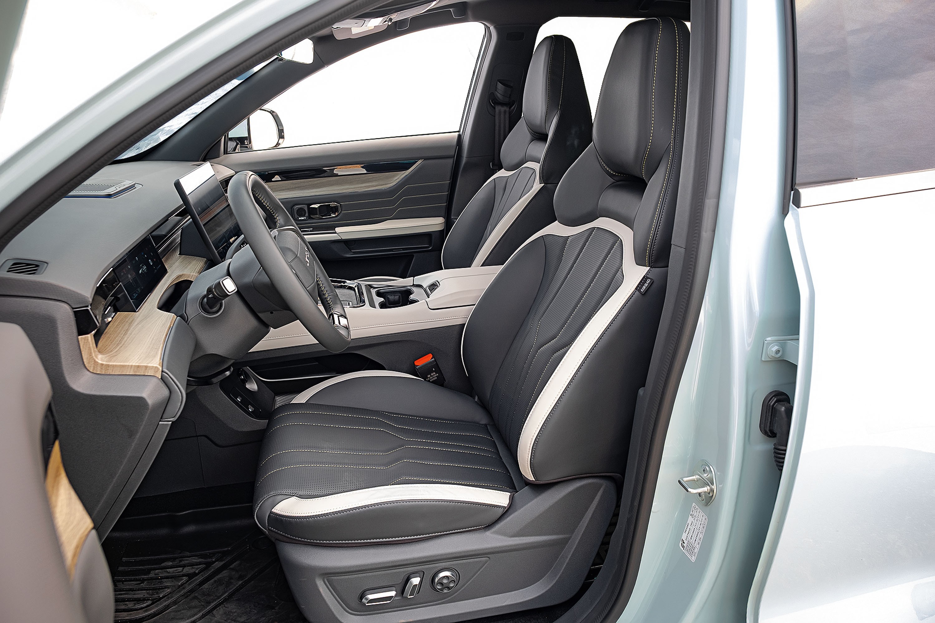 Interior de um carro moderno, com bancos esportivos em couro preto e detalhes brancos, costura aparente. O painel tem acabamento em madeira clara e uma tela digital. O volante é multifuncional, e o console central possui porta-copos. A porta do motorista está aberta, revelando a cor azul-clara da lataria. O ambiente é clean e sofisticado