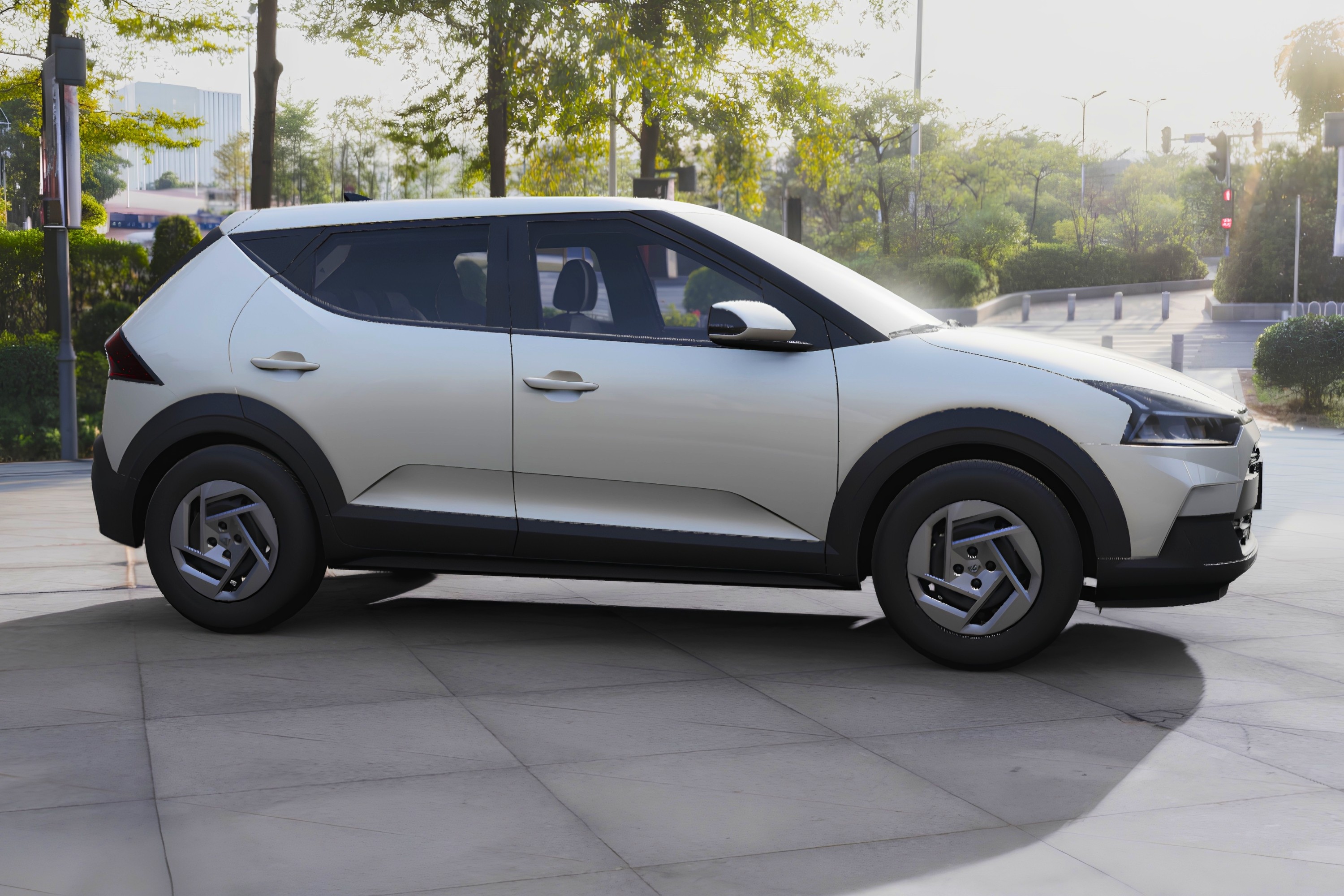 Carro SUV compacto prateado, com detalhes em preto nas caixas de roda e parte inferior, estacionado em calçada de concreto. Ao fundo, árvores e uma rua com semáforo vermelho, sob luz solar