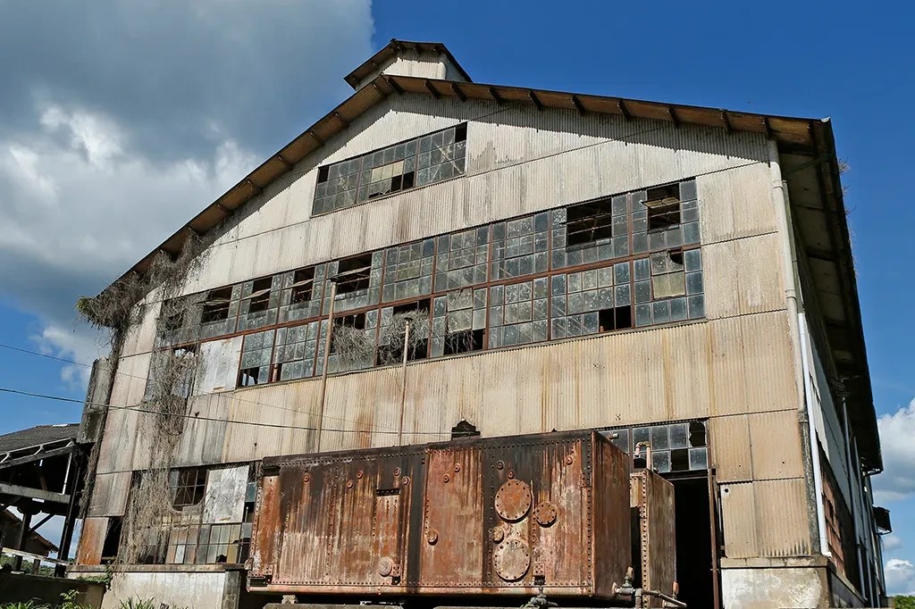 Antiga fábrica abandonada com paredes de chapa metálica, muitas janelas quebradas e vegetação seca subindo pela lateral. Um grande tanque de metal enferrujado está em primeiro plano, sob um céu azul com nuvens brancas