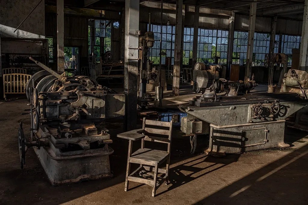 Interior de uma fábrica abandonada, com máquinas industriais antigas e enferrujadas, janelas grandes e sujas, e uma cadeira escolar de madeira no centro, iluminada por um feixe de luz solar que entra pelas janelas e projeta sombras longas no chão empoeirado