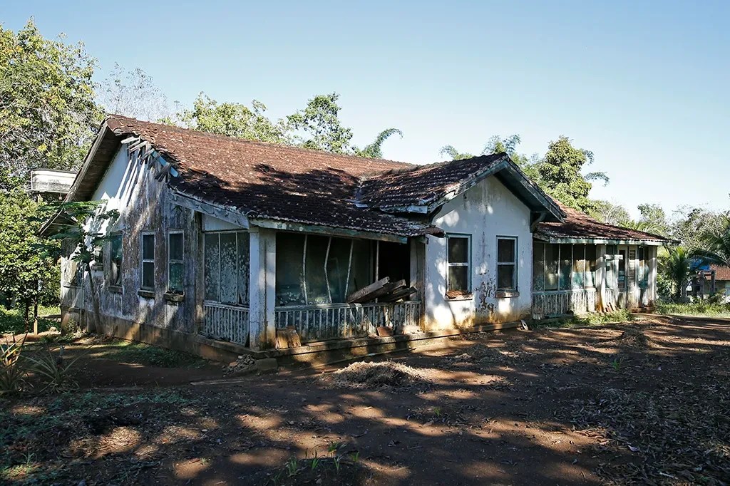 Casa antiga e abandonada, com paredes descascadas, telhado de telhas coloniais marrons e varanda com grades brancas e janelas quebradas. O terreno ao redor é de terra batida com vegetação rasteira e árvores ao fundo, sob céu azul claro