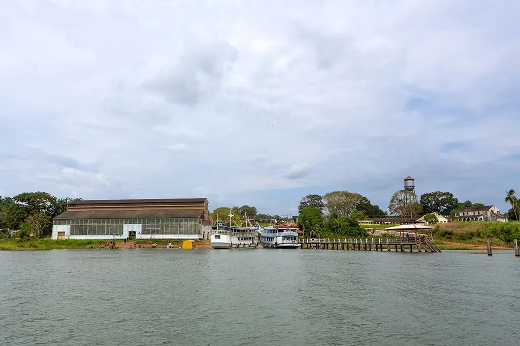 Vista de um rio com margem arborizada, um grande galpão industrial à esquerda, dois barcos atracados, um píer de madeira e um castelo d'água ao fundo, sob céu nublado