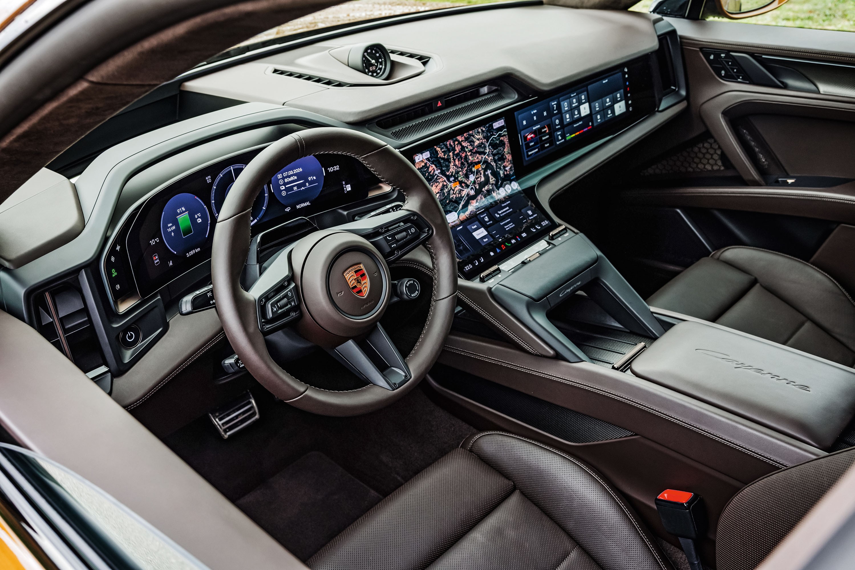 Interior de um carro de luxo com volante marrom escuro e o emblema da Porsche ao centro. O painel possui múltiplas telas digitais exibindo informações do veículo e um mapa. Os bancos são de couro marrom, e o console central tem acabamento sofisticado