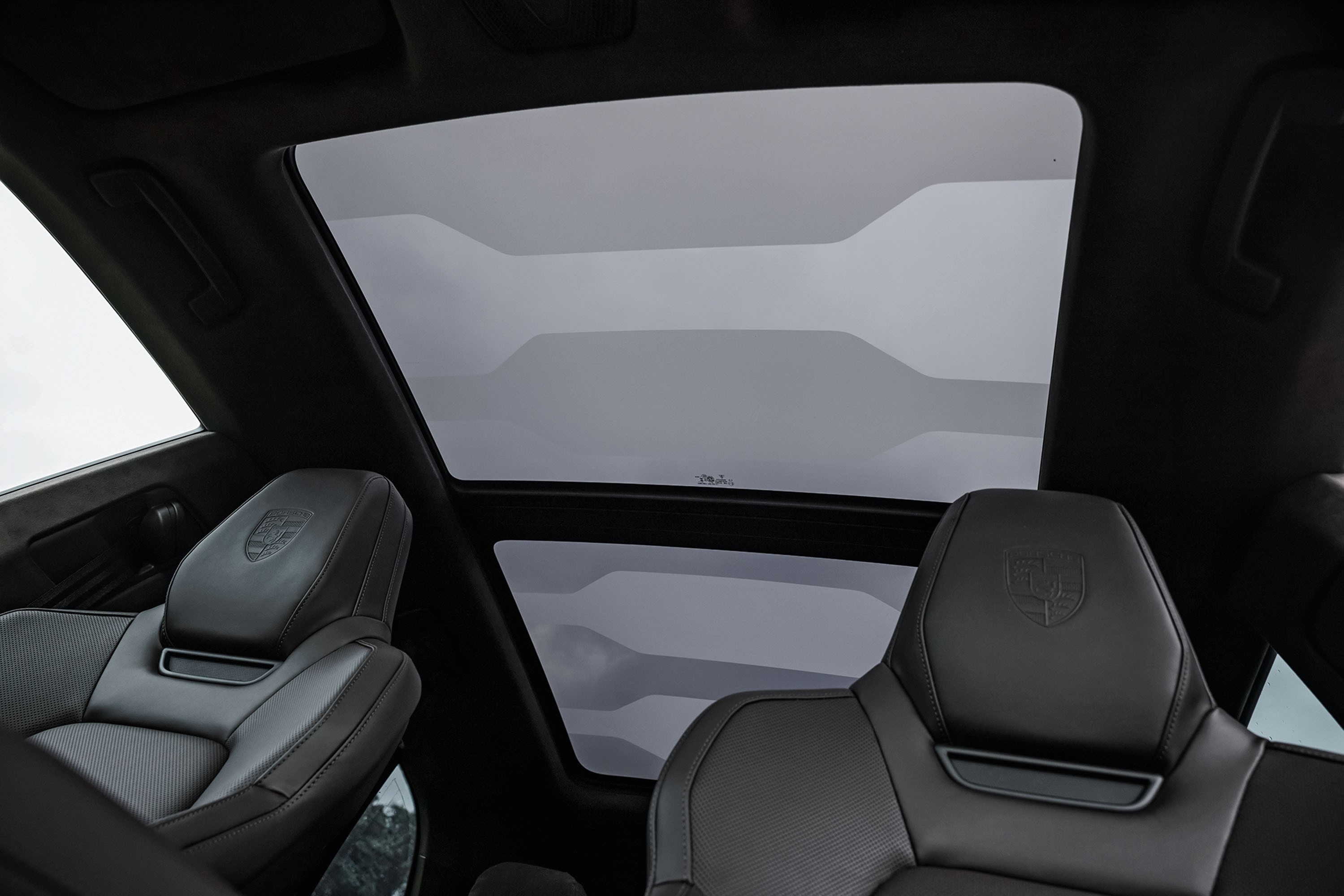 Interior de um carro com teto solar panorâmico e bancos de couro pretos. O teto solar é dividido em duas seções, mostrando um céu cinzento com nuvens. Os encostos de cabeça dos bancos dianteiros exibem um emblema em relevo