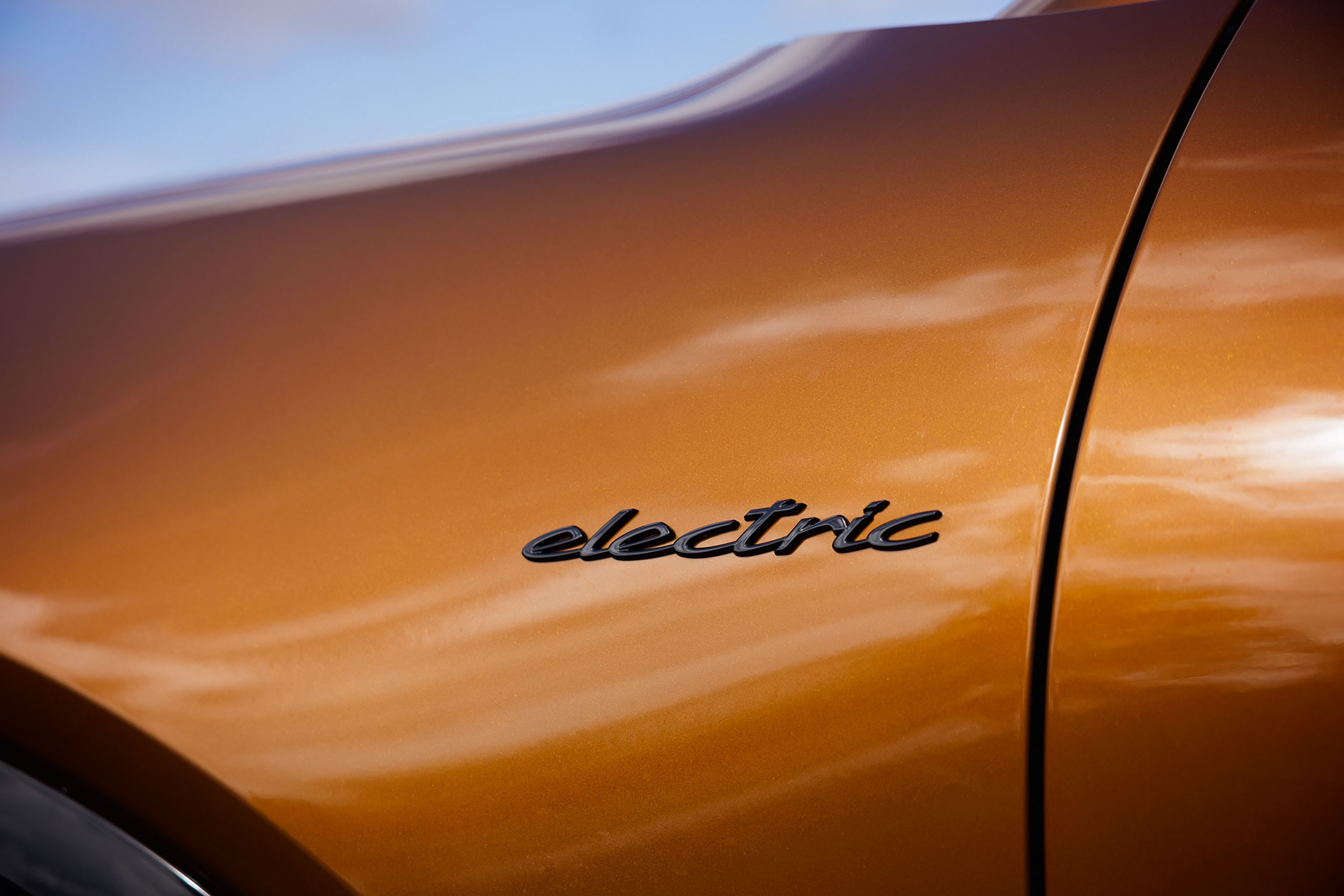 Detalhe da lateral de um carro laranja metálico com o emblema electric em preto, refletindo o céu azul e nuvens