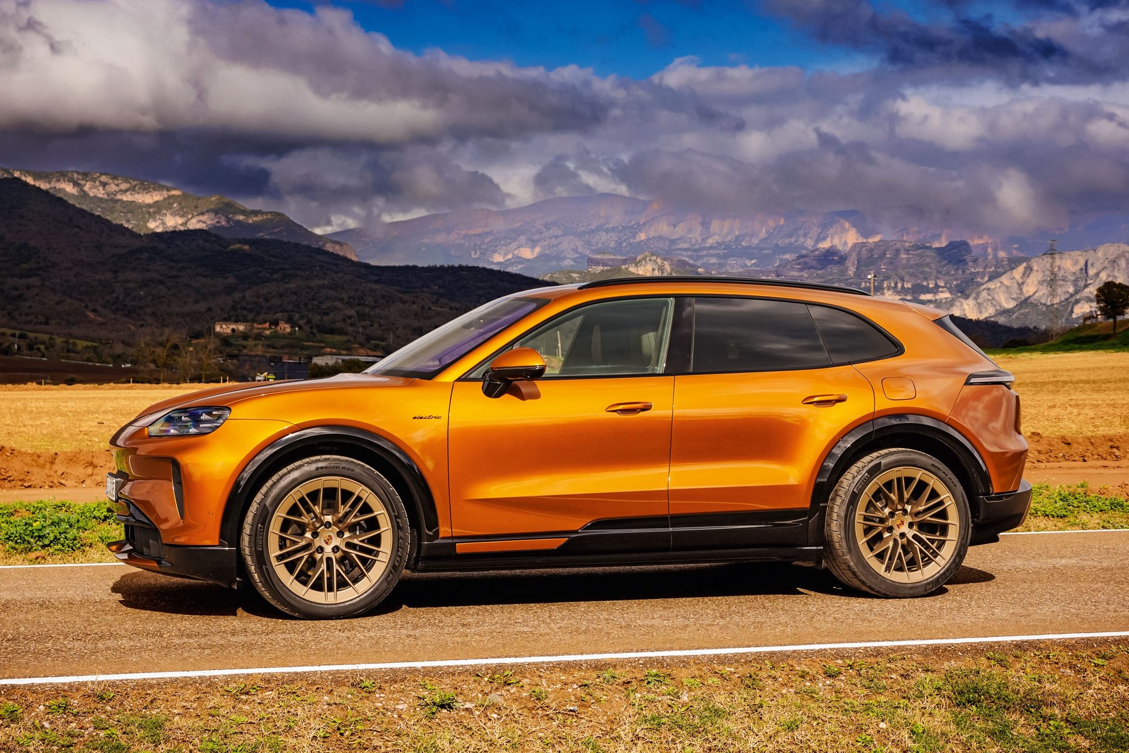 SUV laranja metálico com rodas douradas, parado em uma estrada rural. Ao fundo, montanhas escuras e um céu dramático com nuvens cinzas e trechos azuis. A paisagem tem campos secos e vegetação rasteira