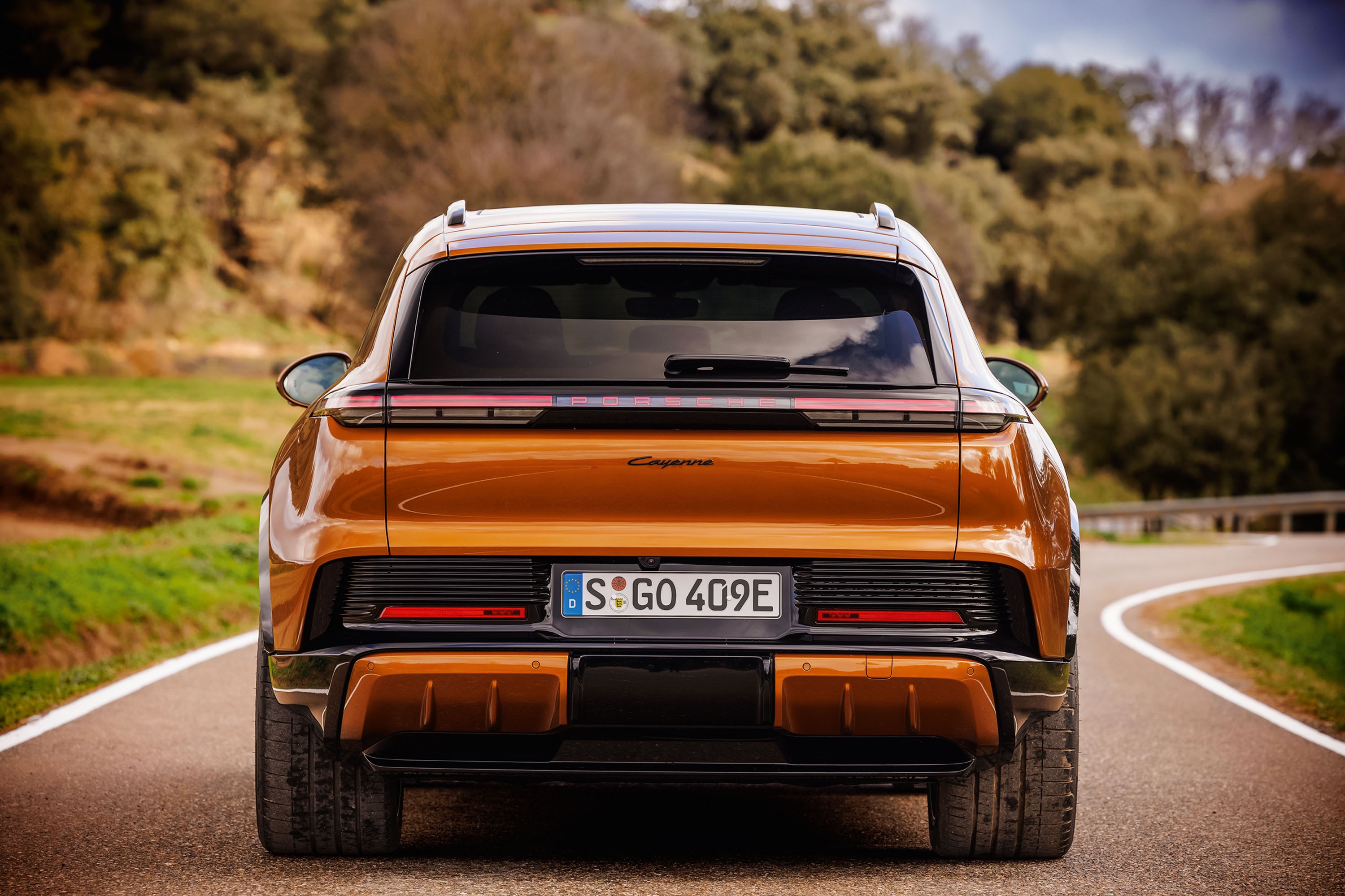 Traseira de um carro SUV Porsche Cayenne laranja metálico, estacionado em uma estrada asfaltada com curvas, cercada por vegetação e árvores