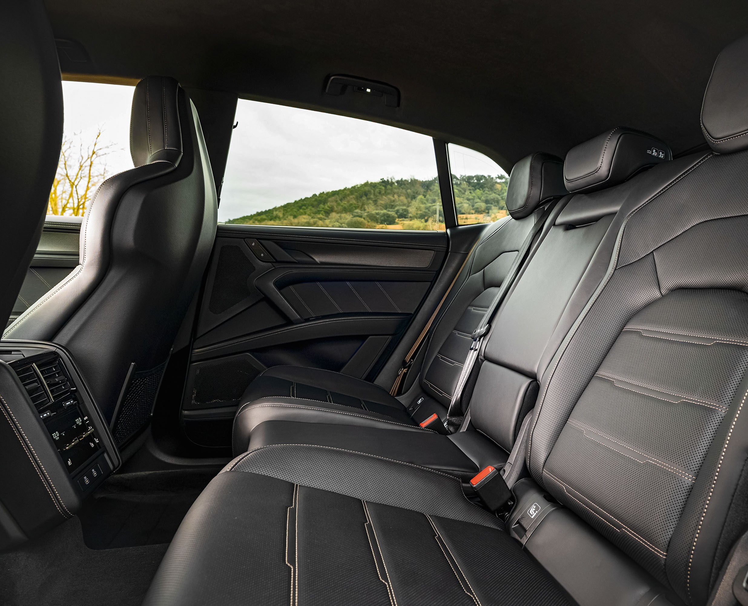 Interior de um carro com bancos traseiros de couro preto perfurado e costuras claras, com encostos de cabeça integrados. Ao fundo, uma paisagem com colinas verdes sob céu nublado, vista pela janela lateral. À esquerda, parte do banco dianteiro e painel com saídas de ar e controles.