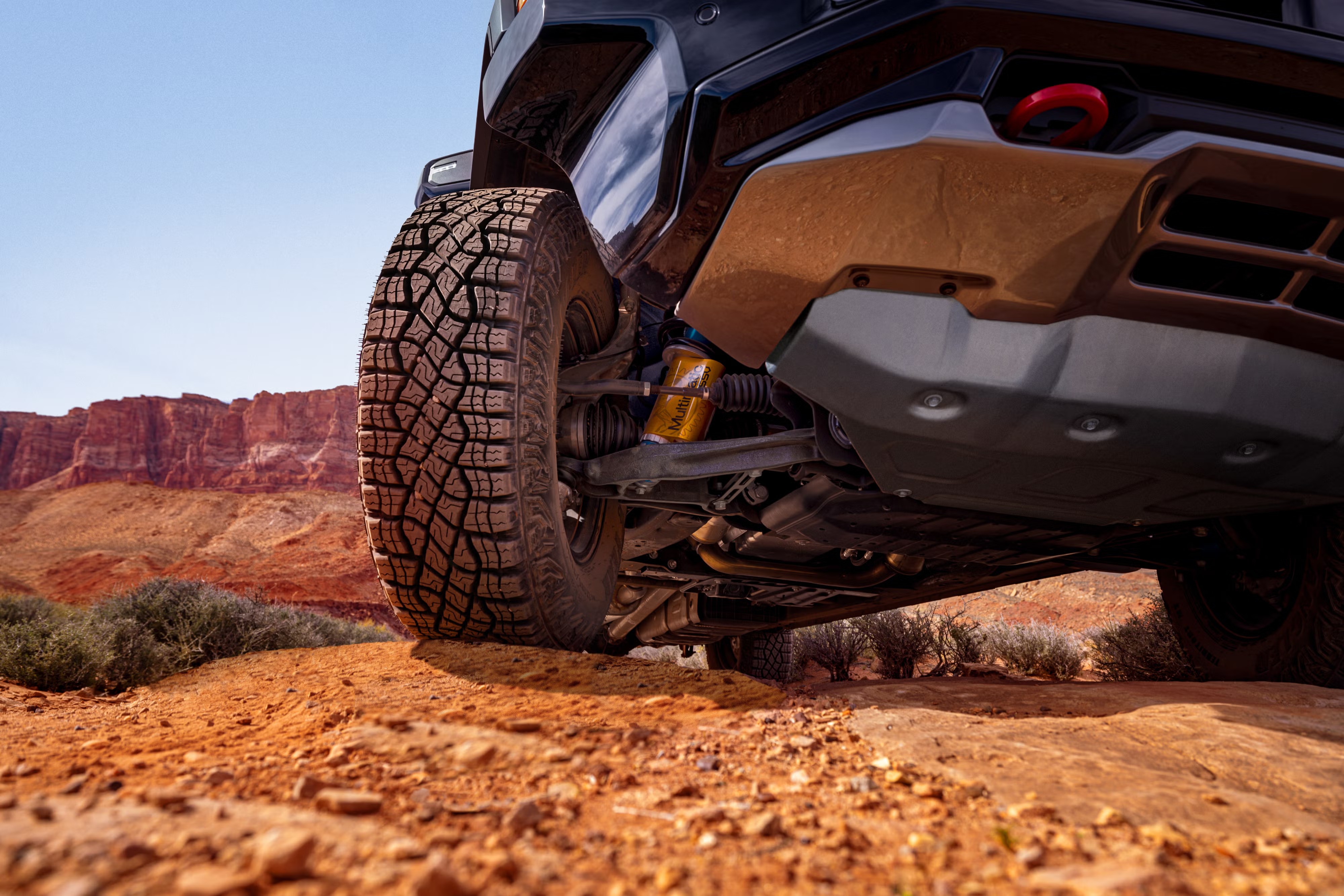 Pneu robusto de um veículo off-road em primeiro plano, sobre terra avermelhada e pedras. Ao fundo, montanhas rochosas e céu azul claro. A suspensão amarela e a parte inferior do carro são visíveis, indicando preparo para terrenos acidentados