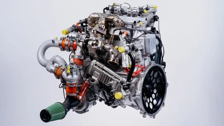 Motor automotivo complexo, com tubulações cinzas e laranjas, detalhes metálicos e um filtro de ar verde na parte inferior esquerda, em fundo branco