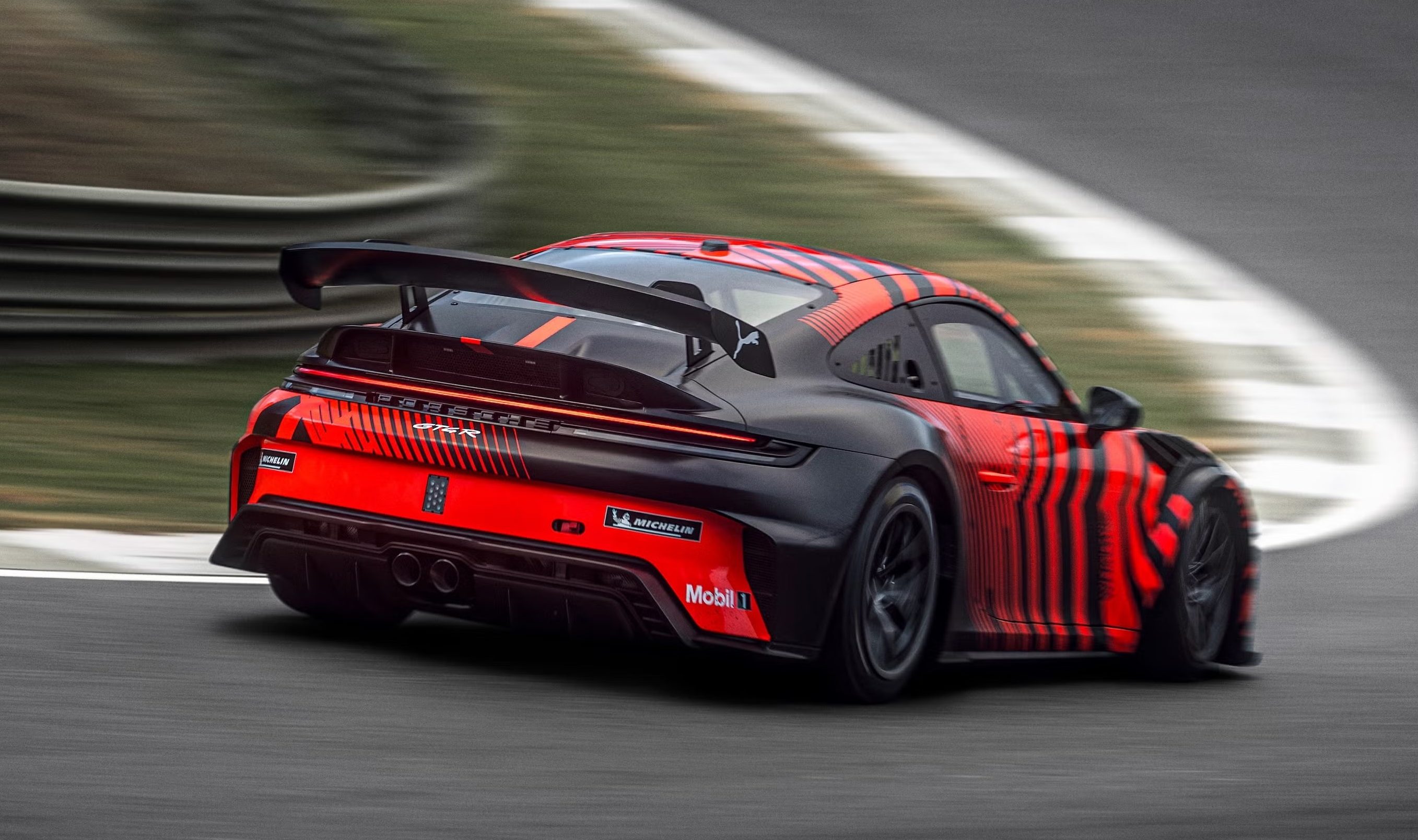 Carro esportivo preto e vermelho com listras verticais, visto de traseira e lateral, em alta velocidade numa pista de corrida cinza, com grama e guard-rail ao fundo