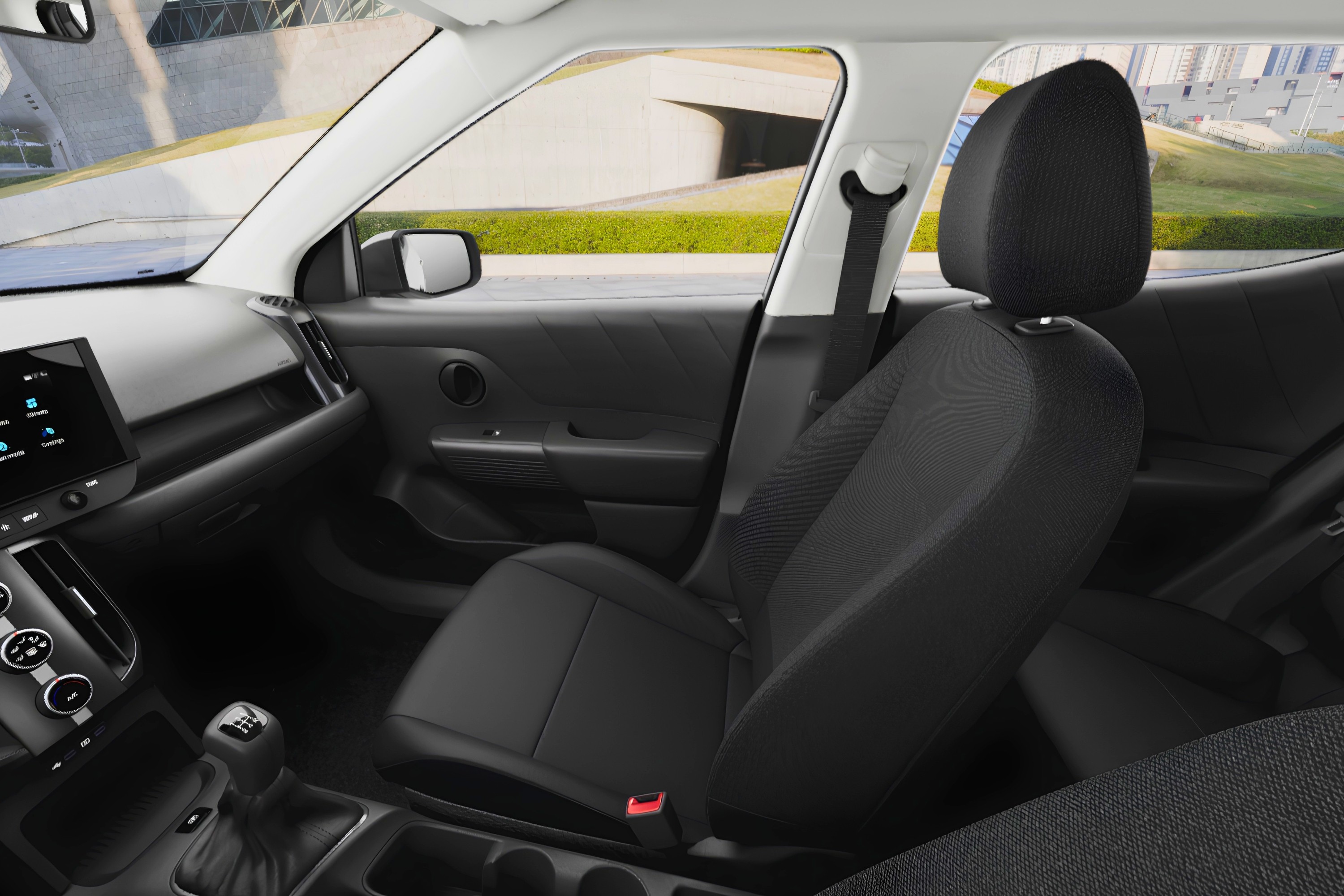 Interior de um carro, mostrando o banco do passageiro dianteiro em tecido preto, o painel com tela multimídia e câmbio manual. Ao fundo, uma estrada com gramado verde e edifícios modernos