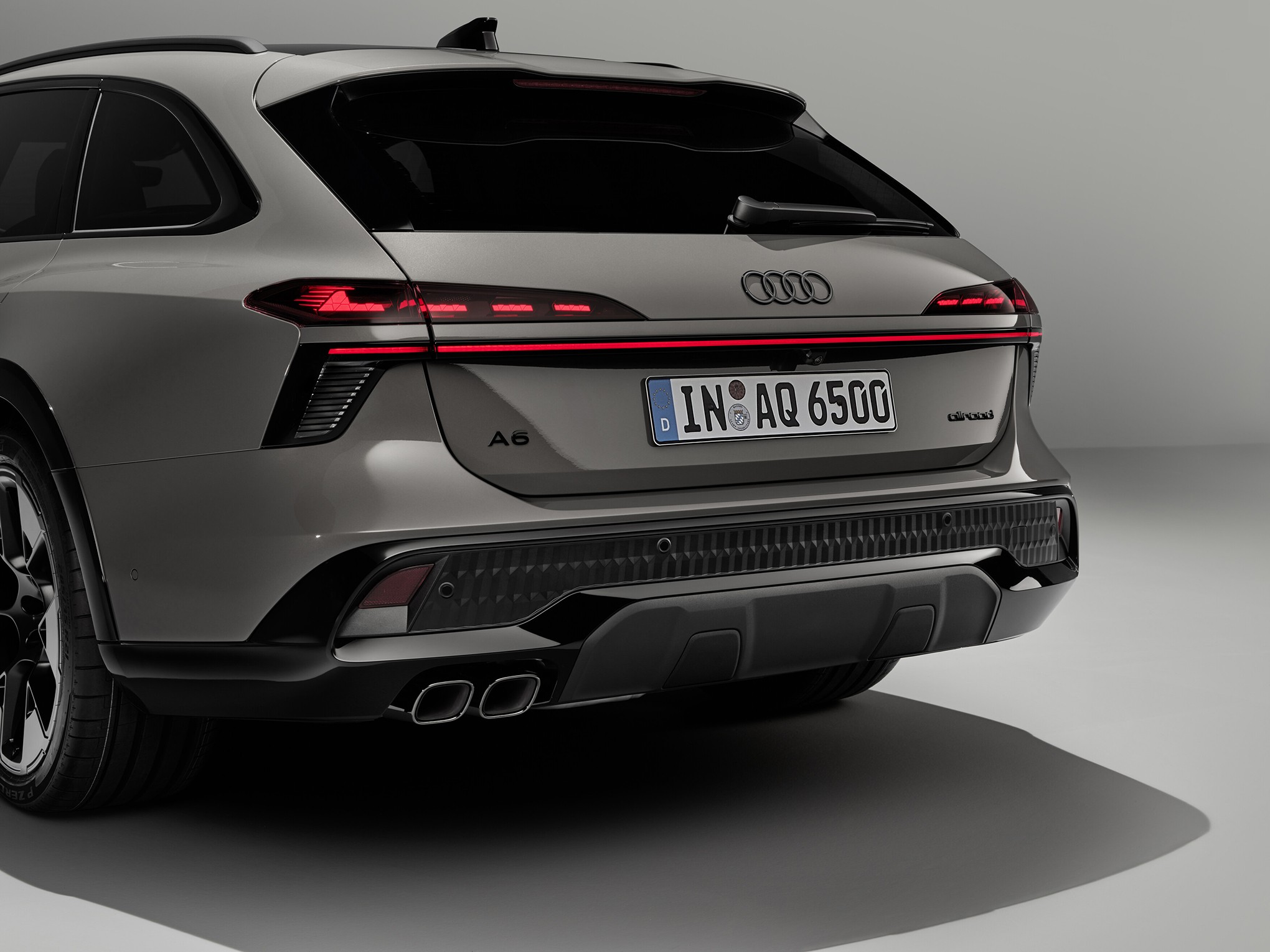 Traseira de um carro Audi A6 cinza-chumbo, com lanternas vermelhas acesas e uma faixa de LED horizontal. A placa de licença alemã exibe IN AQ 6500. O para-choque traseiro tem detalhes pretos e duas saídas de escapamento cromadas. O carro está estacionado em um ambiente claro, com uma sombra projetada no chão