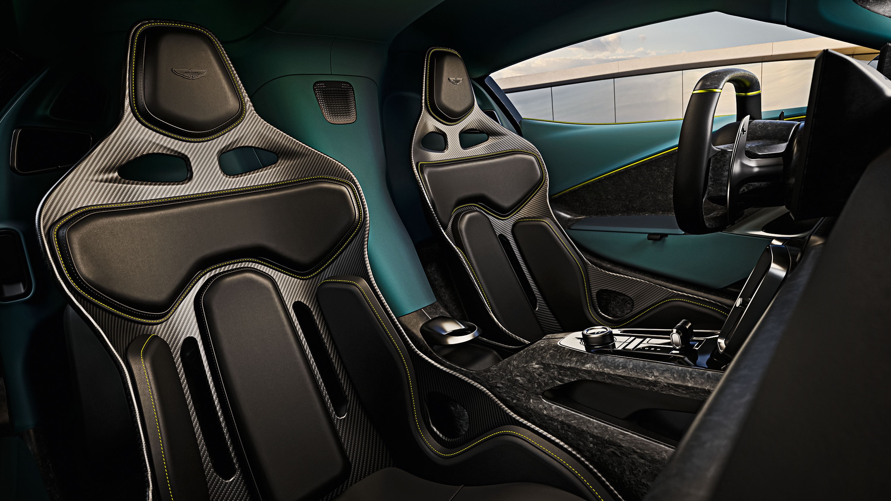 Interior de carro esportivo com dois bancos concha pretos e cinzas de fibra de carbono, costura amarela. Painel e portas em tom verde-azulado, volante à direita. Console central com detalhes em fibra de carbono e botões prateados. Janela lateral exibe um céu claro e uma parede cinza ao fundo