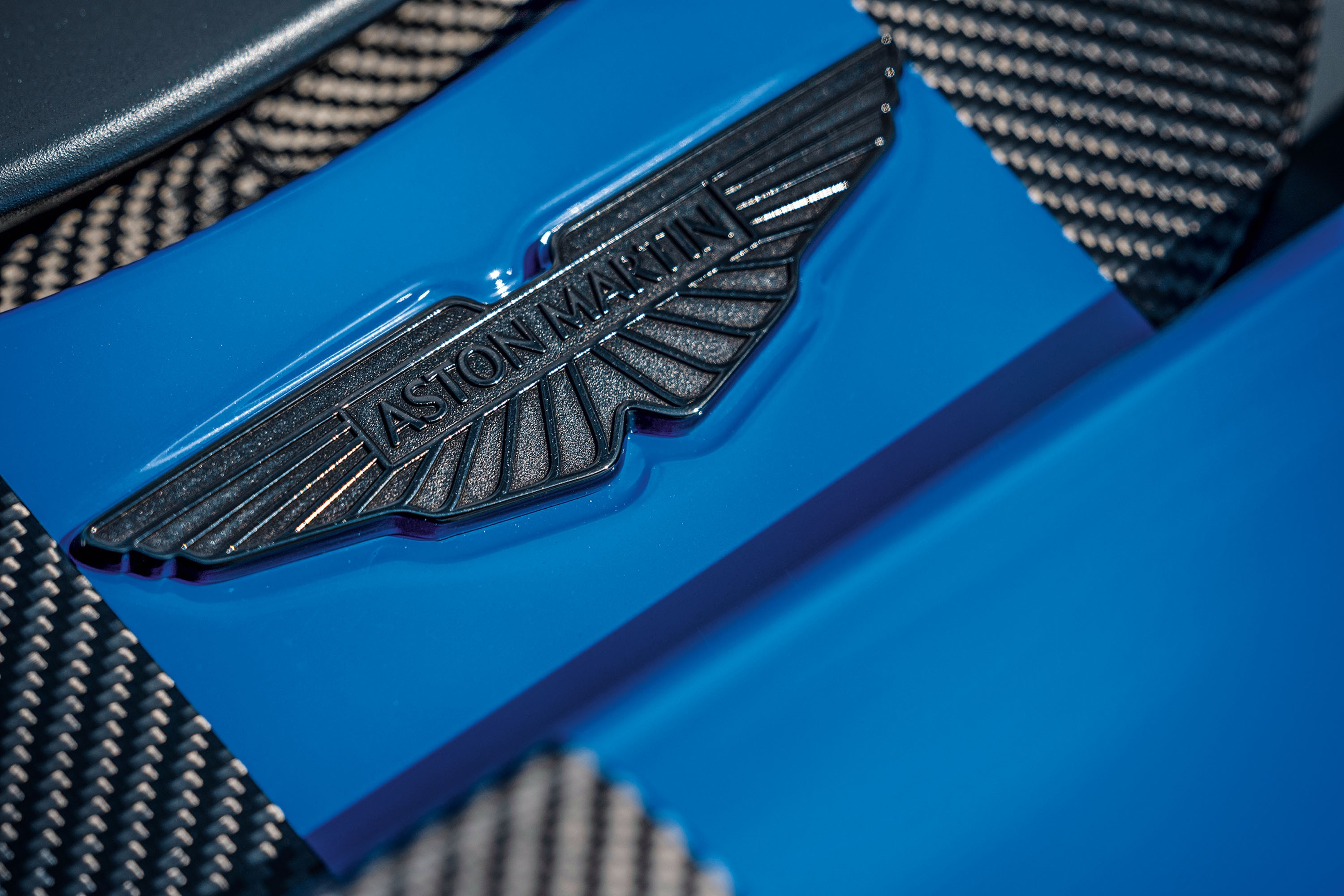 Emblema metálico cinza escuro da Aston Martin com asas e o nome da marca em relevo, sobre uma superfície azul brilhante e um tecido quadriculado cinza e branco ao fundo