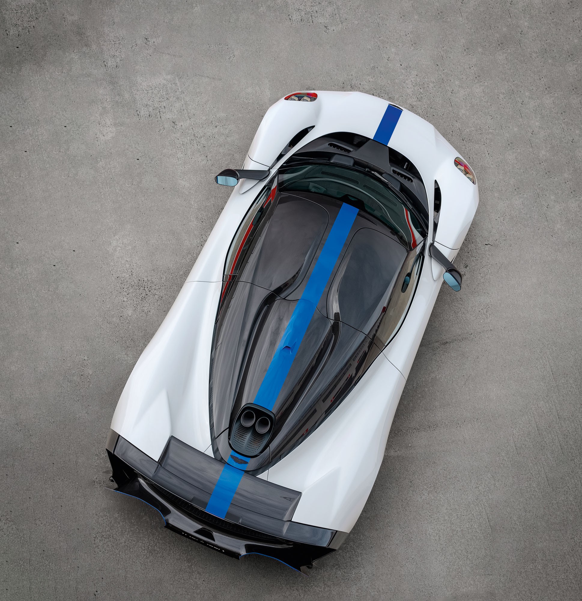 Carro esportivo branco com faixa azul central, visto de cima, estacionado em piso de concreto cinza. O teto é de vidro escuro, e a traseira possui duas saídas de escapamento redondas