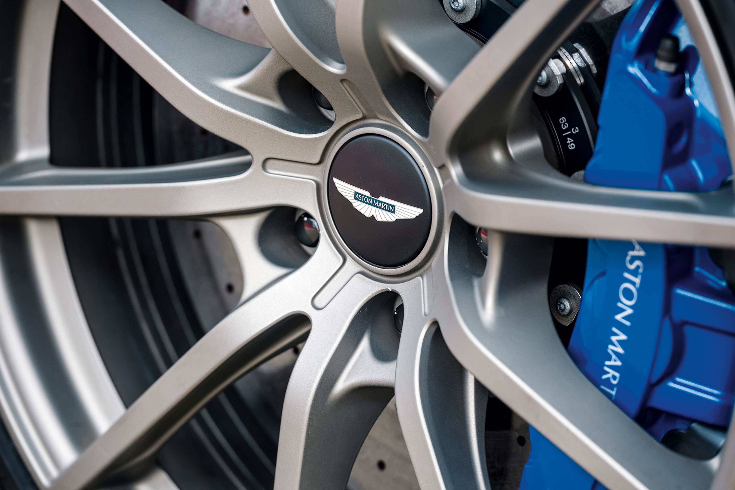 Roda de carro esportivo Aston Martin com aro cinza-chumbo de múltiplos raios, pinça de freio azul brilhante e o logotipo da marca no centro