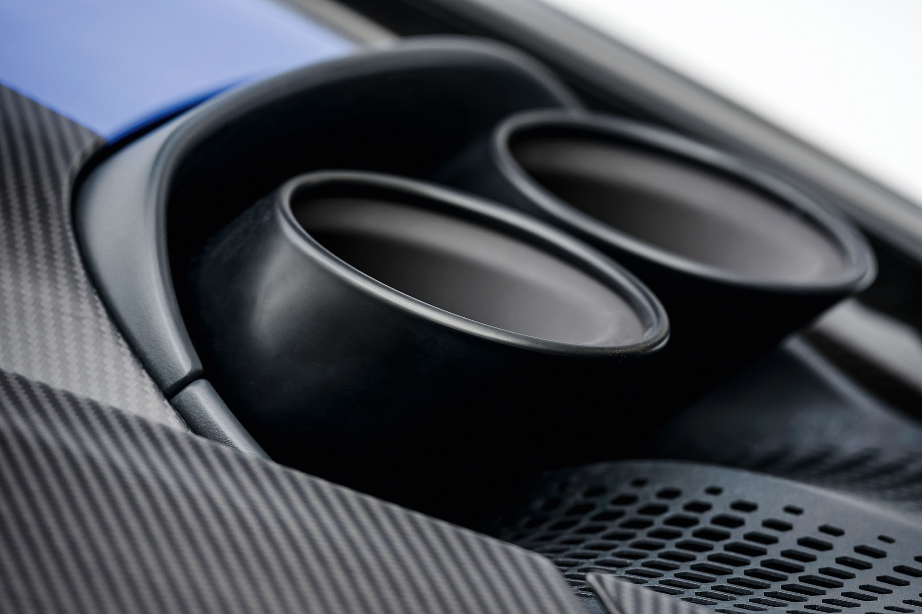Detalhe da traseira de um carro esportivo azul, mostrando duas saídas de escapamento pretas foscas e parte da carroceria em fibra de carbono com textura diagonal. Abaixo dos escapamentos, uma grade preta com padrão hexagonal.