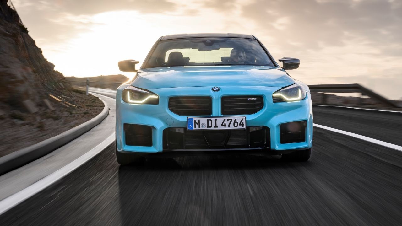Carro esportivo BMW M2 azul claro em uma estrada sinuosa, visto de frente, com faróis acesos e pôr do sol ao fundo